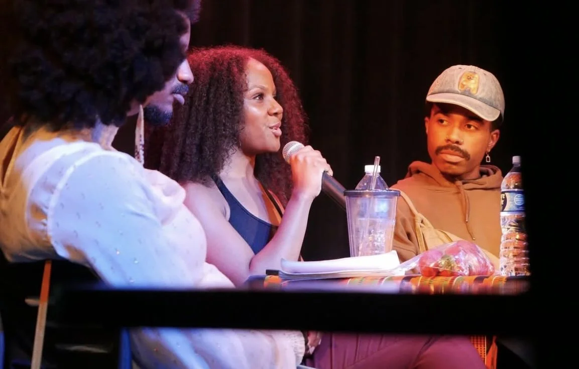Three people sitting at a table on stage, with the woman in the middle speaking into a microphone, while the others listen. The table has drinks, a water bottle, a laptop, and some items on it.