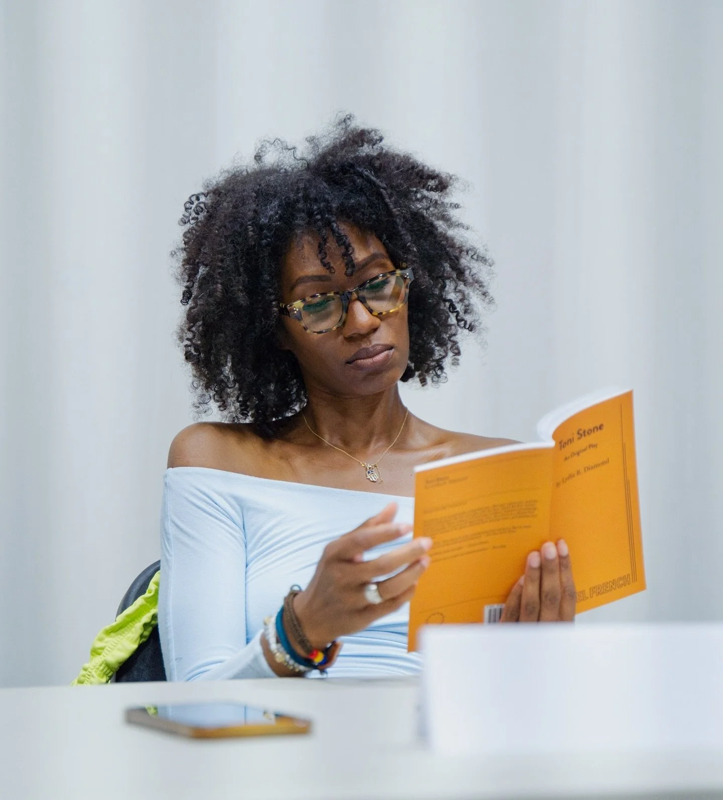 A woman with curly hair, glasses, and bracelets, sitting at a table, reading an orange book.