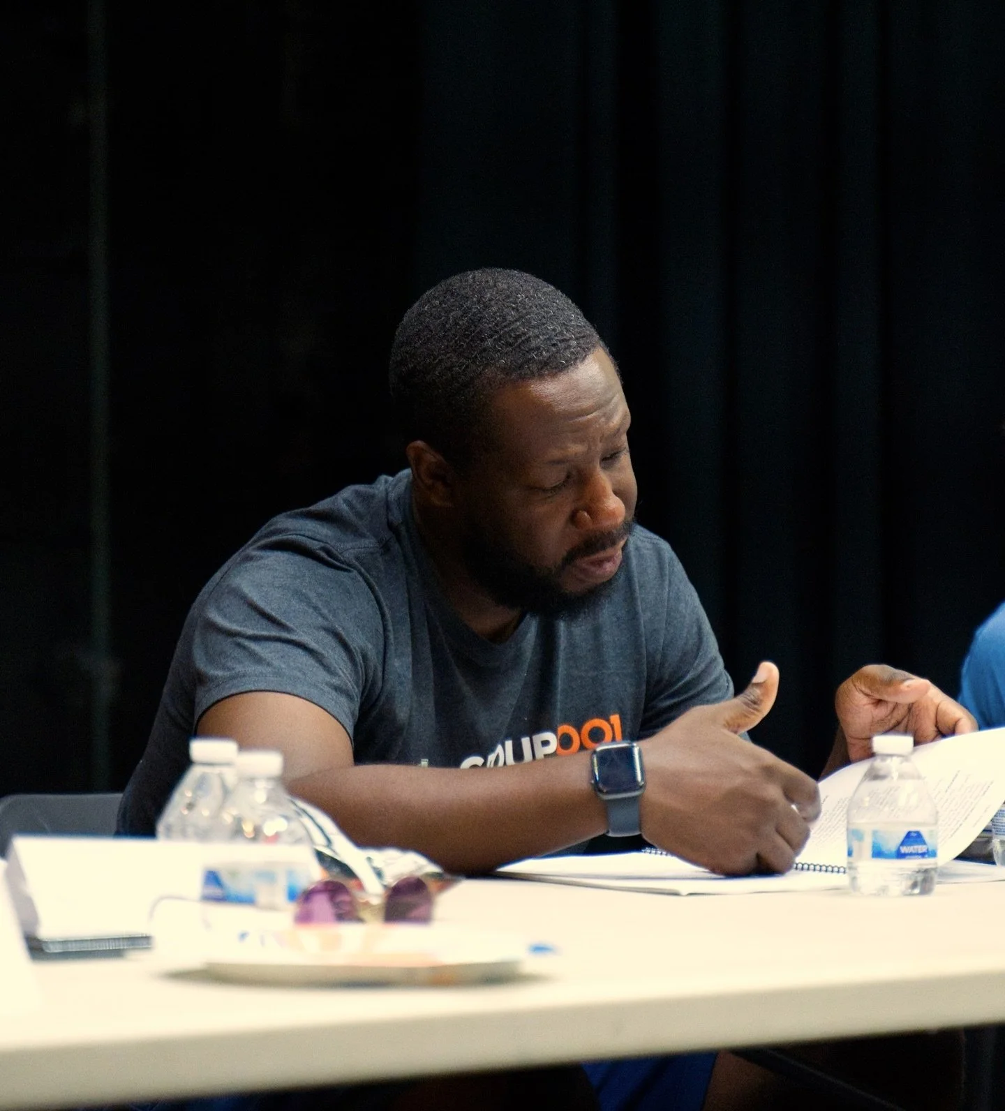 A man with a beard wearing a dark t-shirt and smartwatch sitting at a table, reading a document with water bottles and glasses on the table.