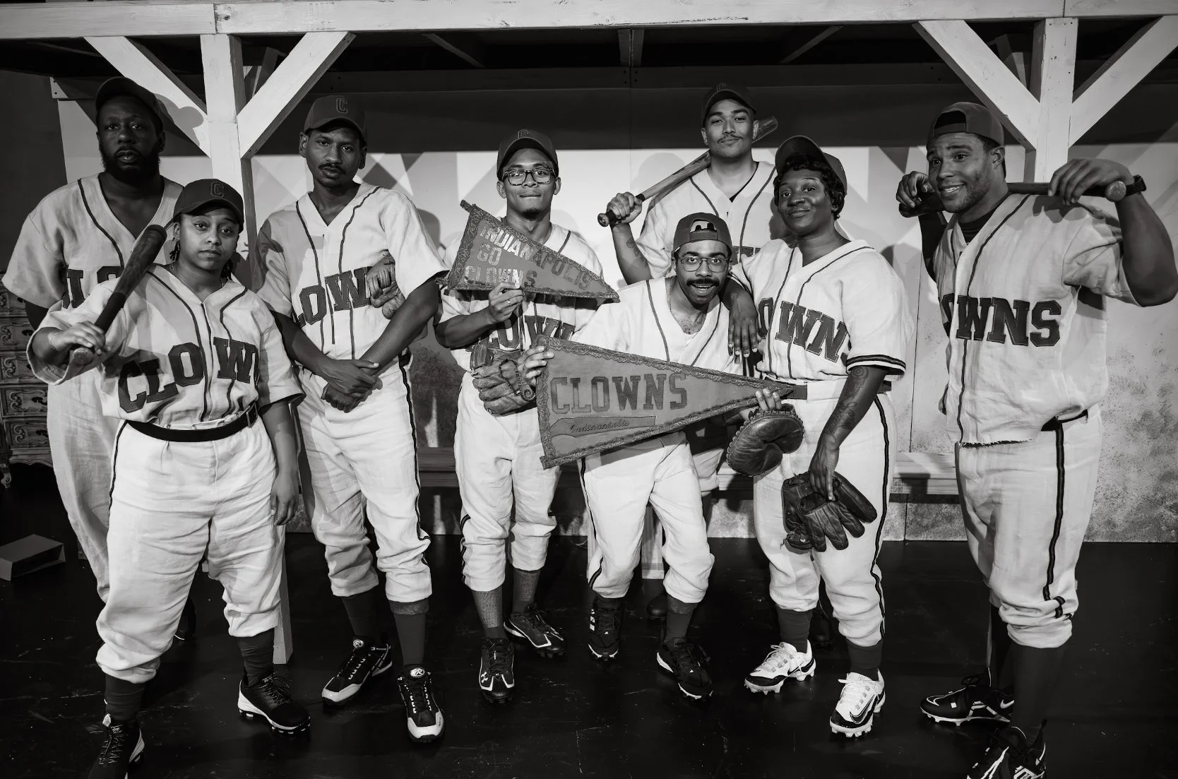 A group of nine baseball players in uniform posing indoors, holding baseball bats and flags, with 'Clowns' written on some banners, celebrating a team victory.