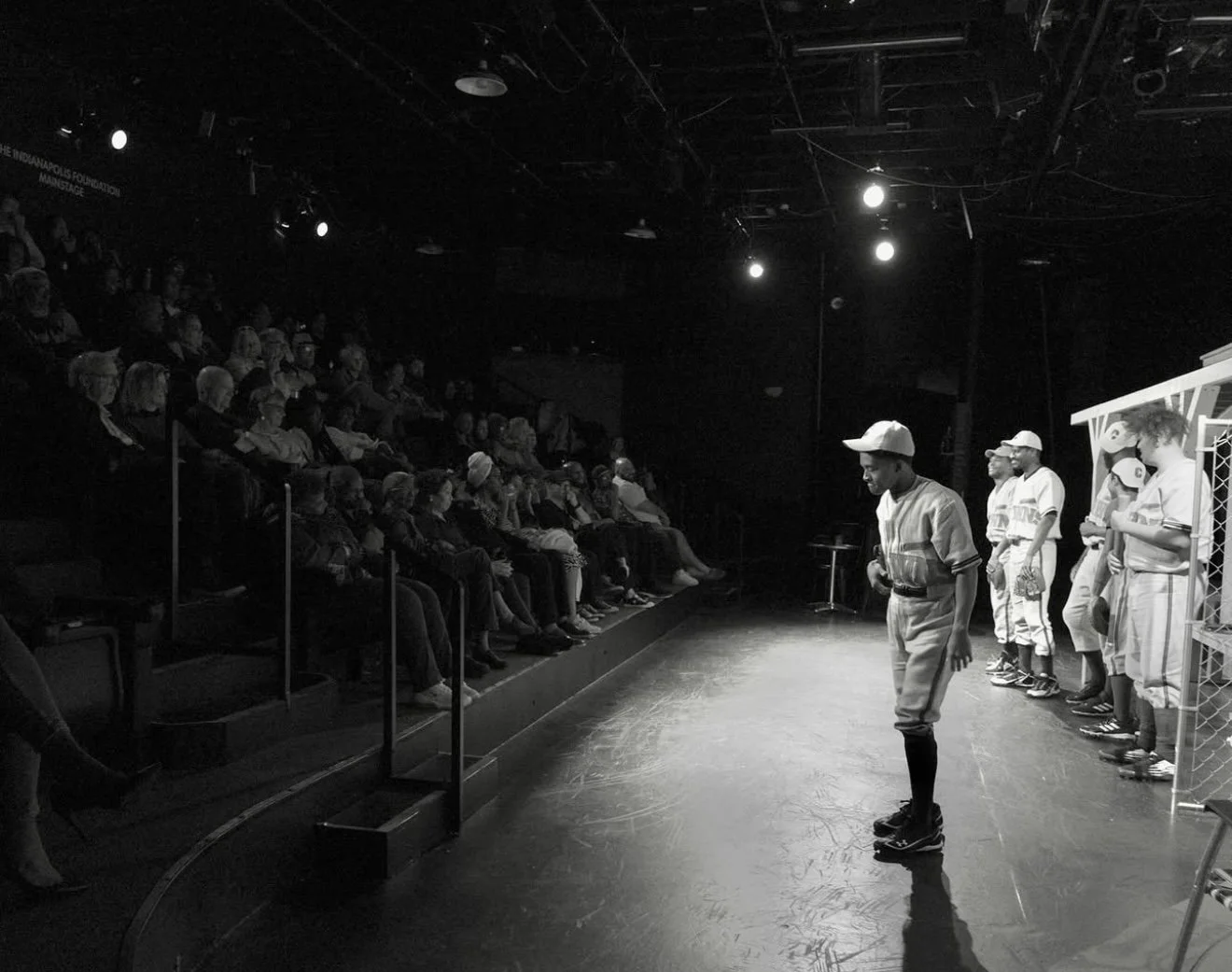 A black and white photo of a theatrical performance. A person in a baseball uniform is on stage, bending forward, while other performers in baseball uniforms stand behind him. The audience is seated on the left side, attentively watching the performa