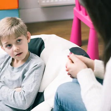 A woman and a girl sitting on the floor in a living room, engaged in a coloring activity. The girl is holding a paper with a sad face emoji, and the woman is writing on a clipboard. There are colored pencils scattered on the floor, and pictures on the wall behind them.