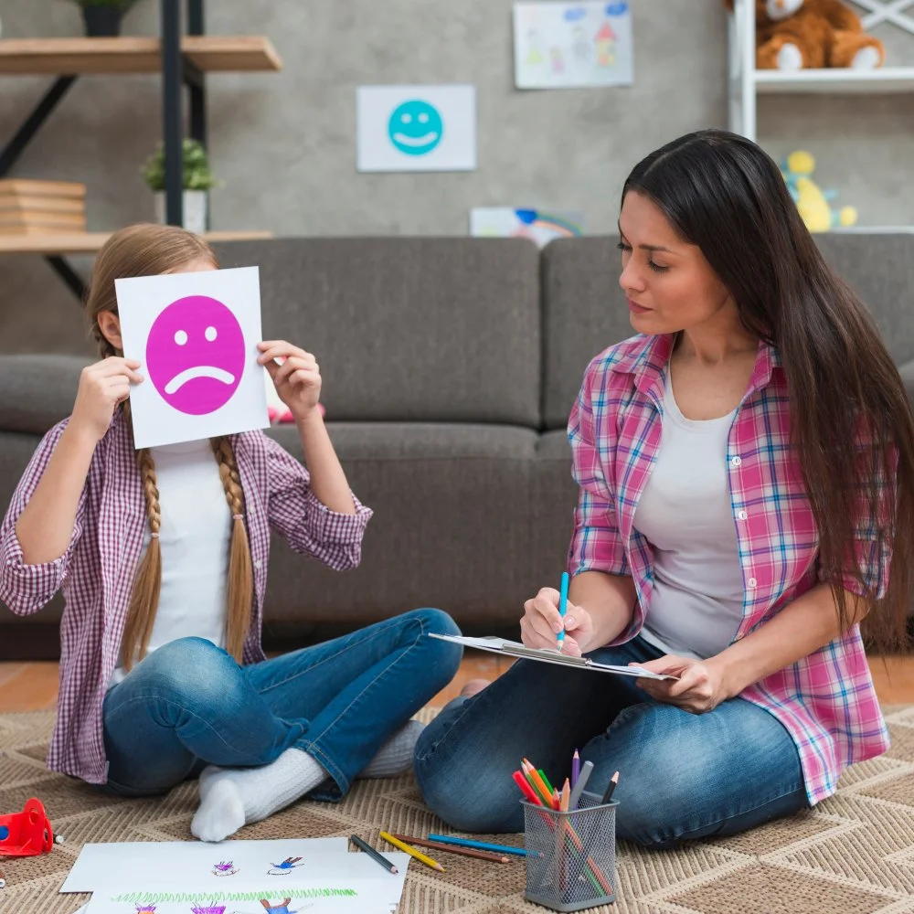 A young girl sitting on the floor holding a paper with a purple sad face mask in front of her face. An adult woman, likely her mother or therapist, sitting nearby with a notepad and pen, observing her. The setting appears to be a cozy living room with art on the wall and books on shelves.
