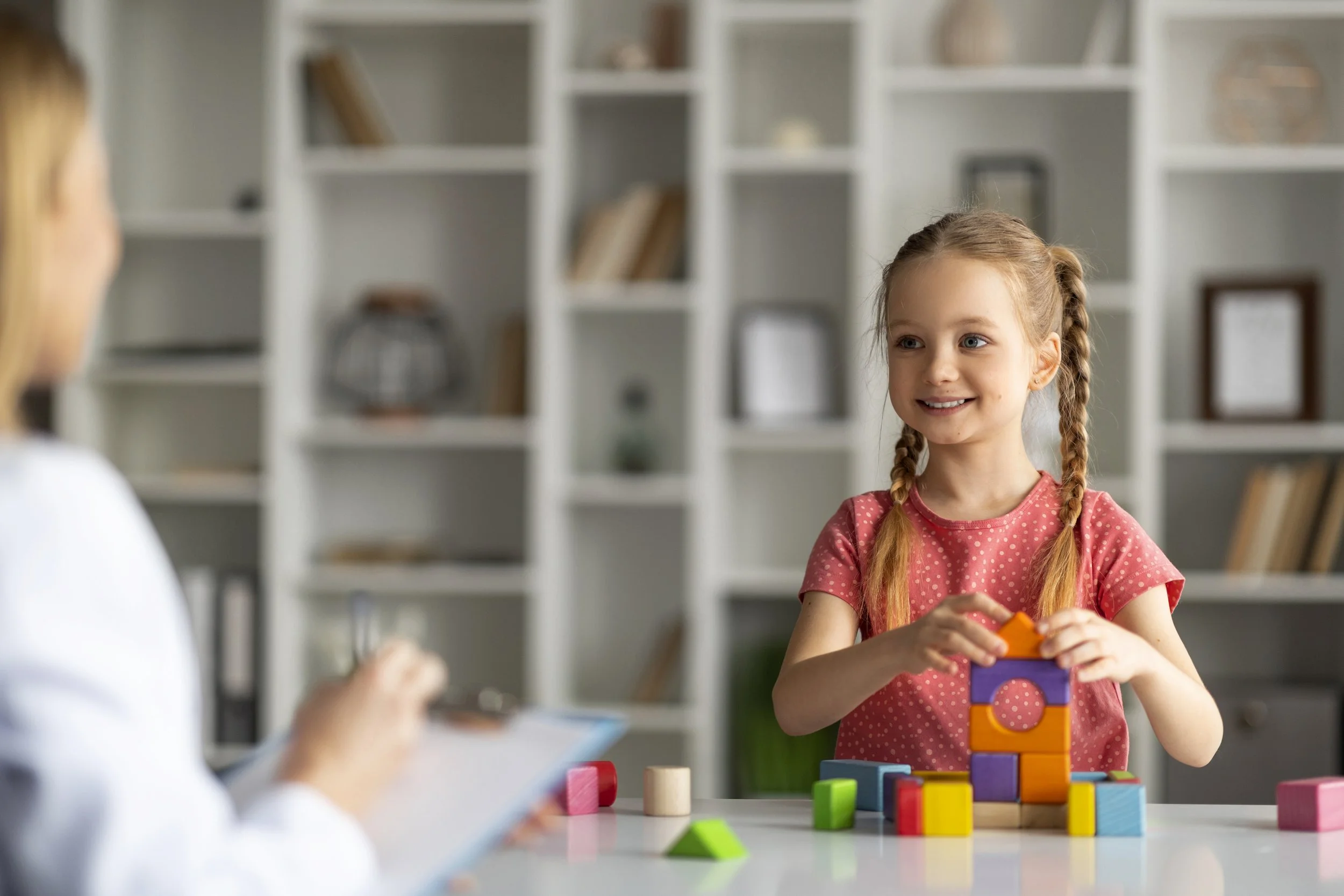 A young girl with braids smiling and stacking colorful blocks at a table in a room with a white bookshelf in the background.