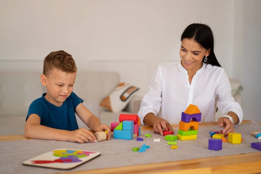 A woman and a young boy playing with colorful wooden building blocks at a table in a living room.