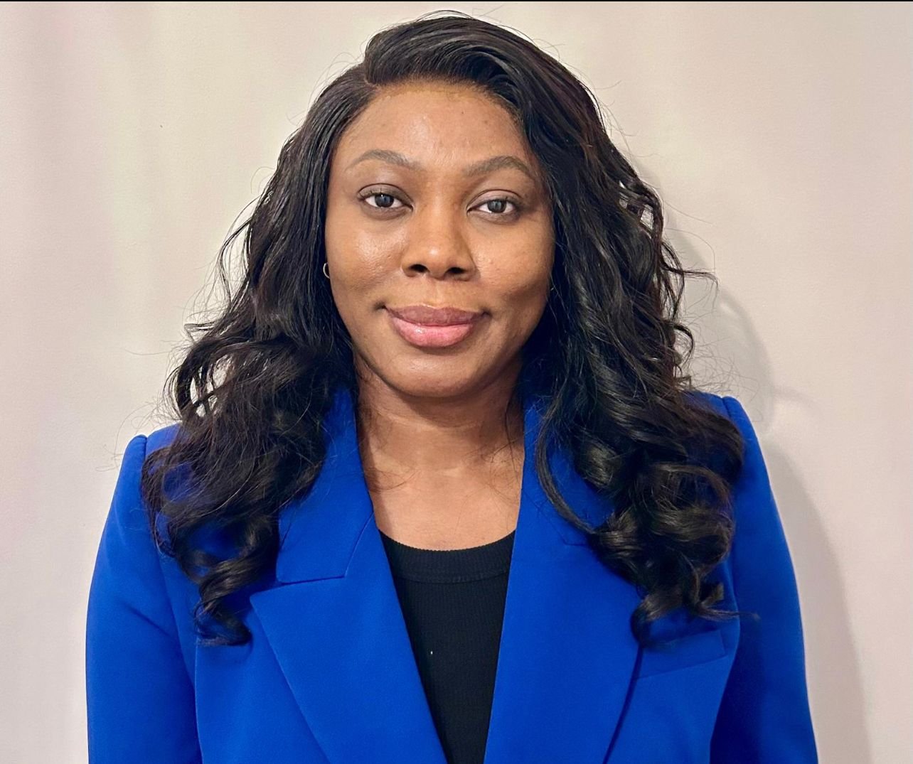 A woman with long, curly black hair wearing a blue blazer and a black top, standing against a plain light-colored wall.