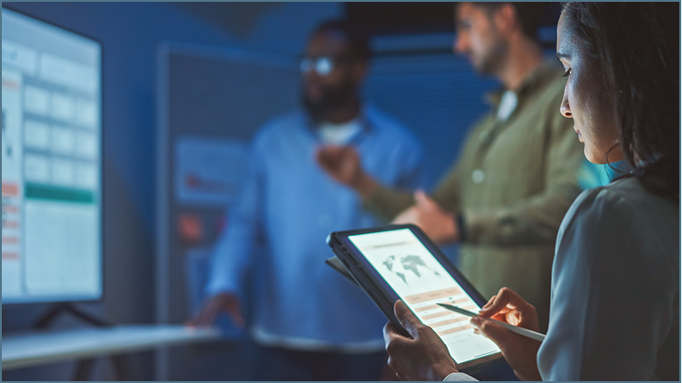 A woman in a business suit holding a tablet and a smartphone in a conference room with three men. The men are standing near a presentation screen, engaged in a discussion.