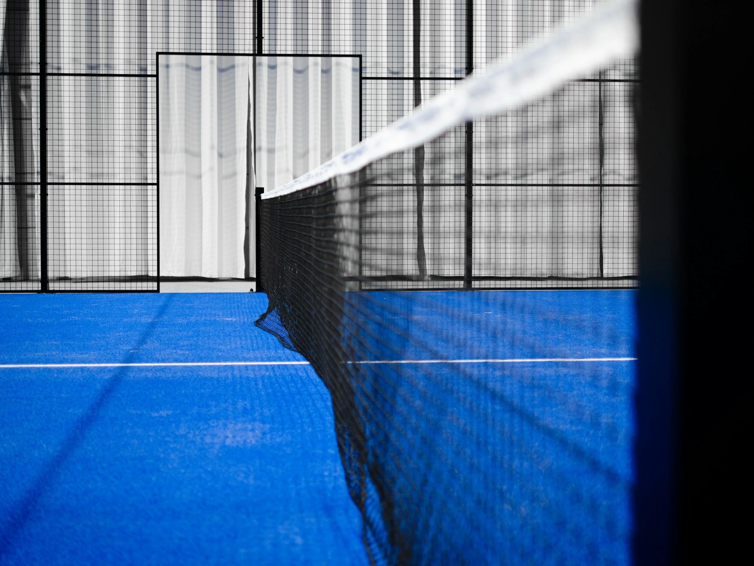 Indoor tennis court with a black net, blue court surface, and white curtains in the background.