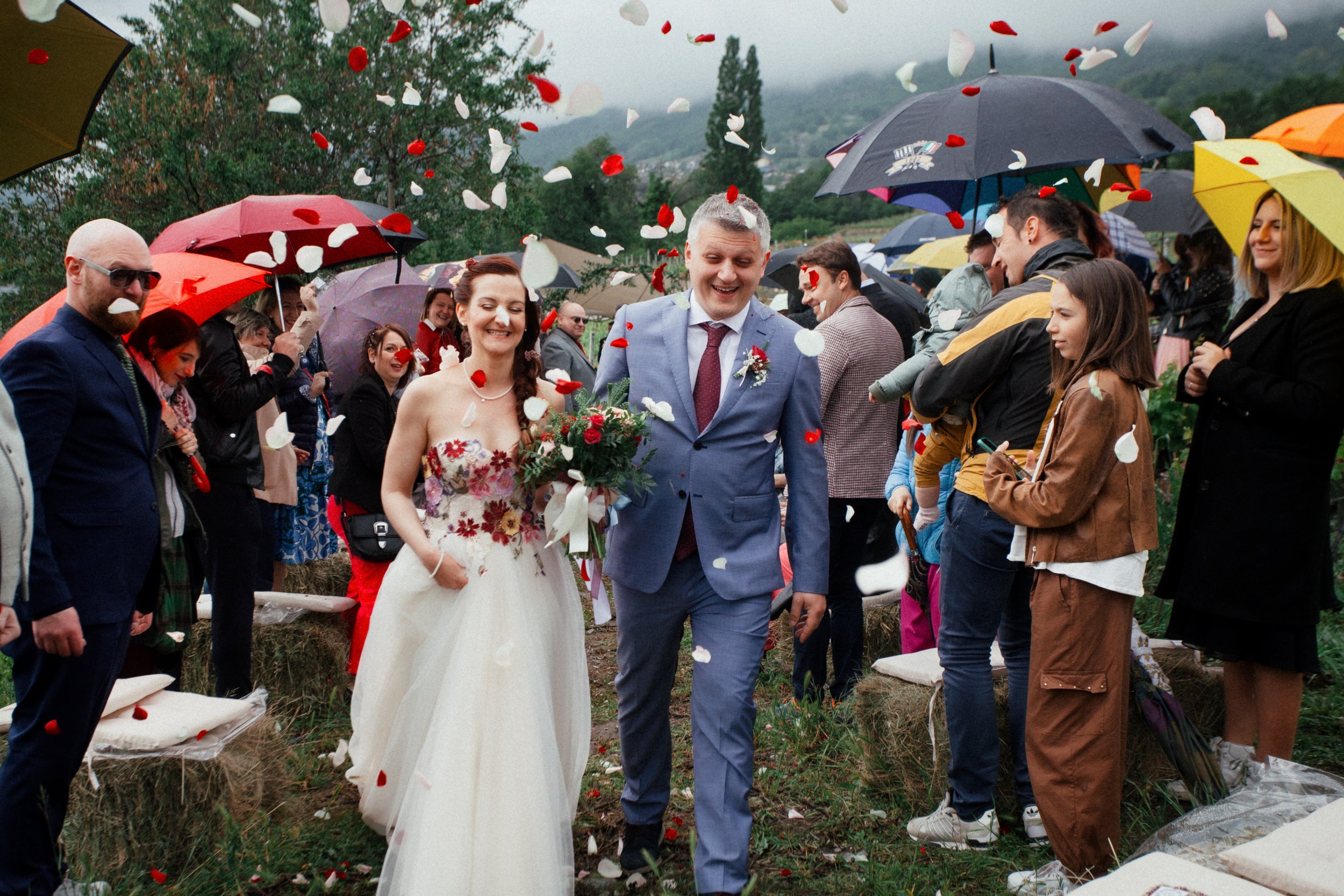 Una coppia di sposi cammina sotto petali di fiori lanciati dagli invitati, alcuni dei quali reggono ombrelli colorati. Sullo sfondo si vedono colline e alberi.