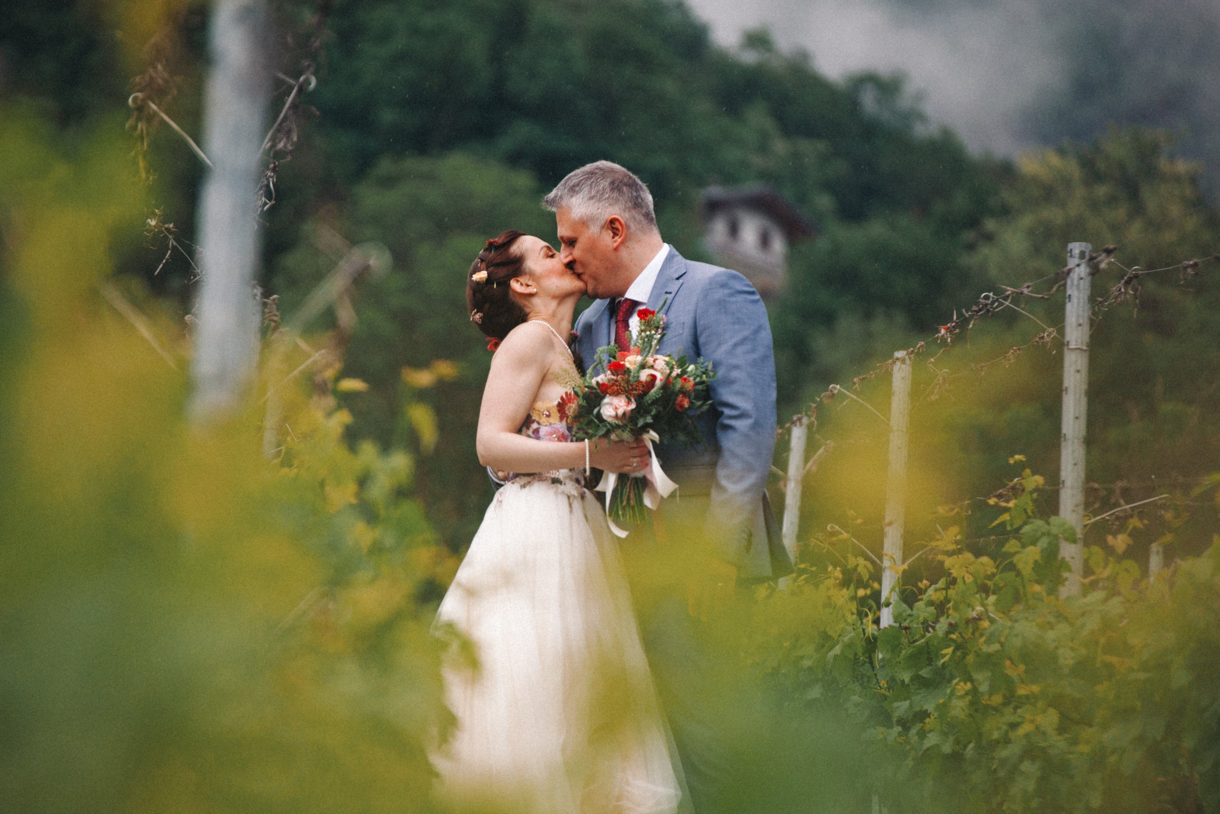 Coppia di sposi che si baciano in un vigneto, con sfondo di alberi e vegetazione. La sposa indossa un abito bianco e tiene un bouquet di fiori, mentre lo sposo è in un abito grigio.