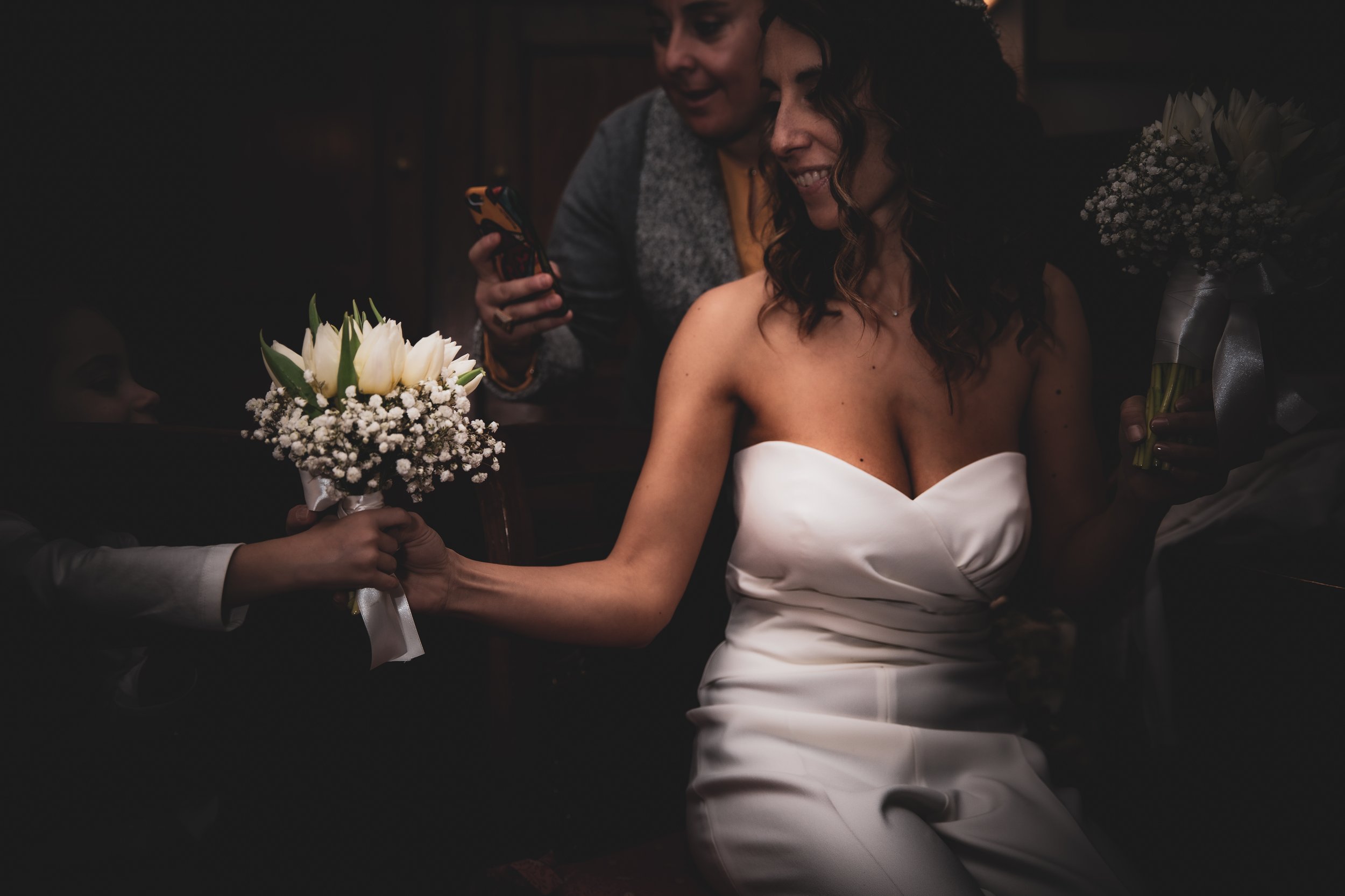 Una donna in abito da sposa bianco sorride mentre porge un bouquet di fiori bianchi a una persona. Una seconda persona tiene un telefono in mano sullo sfondo.