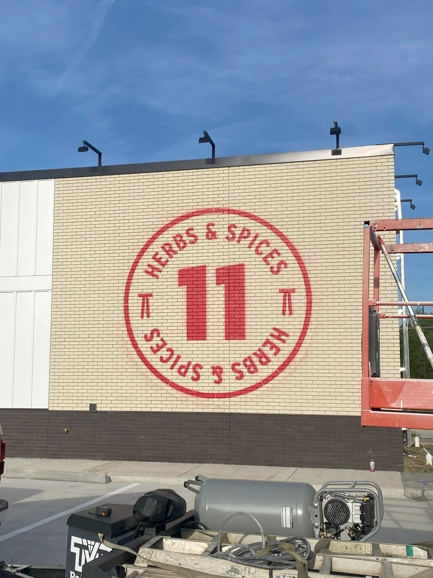 Construction site with a large brick wall painted with the logo for a restaurant, multiple black outdoor lights on top of the wall, and equipment including scaffolding and machinery in the foreground.