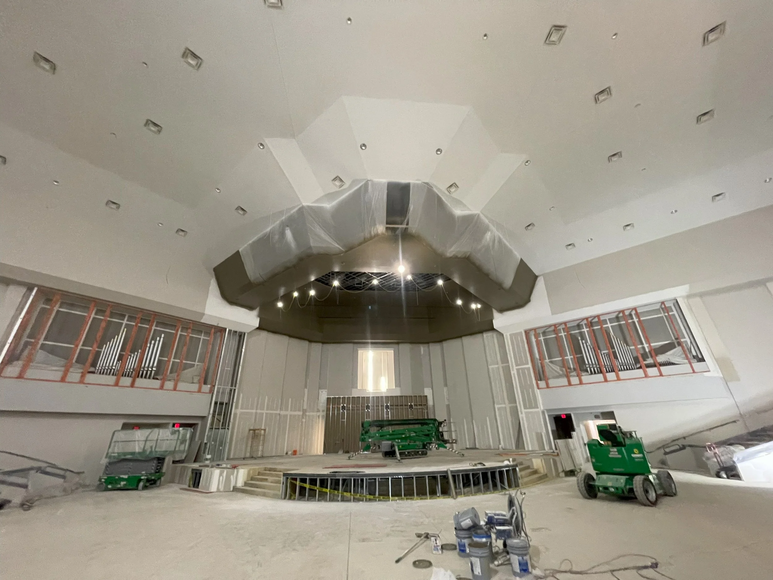 Interior of a building under construction or renovation, with visible ceiling fixtures, construction equipment, and unfinished areas.