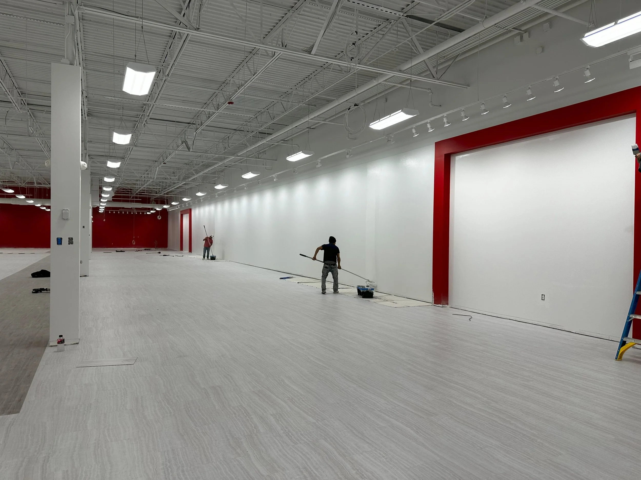 Interior of a large, TJ Maxx showroom under construction, with workers  painting the walls, high ceilings with exposed metal beams, bright ceiling lights, and red and white walls.