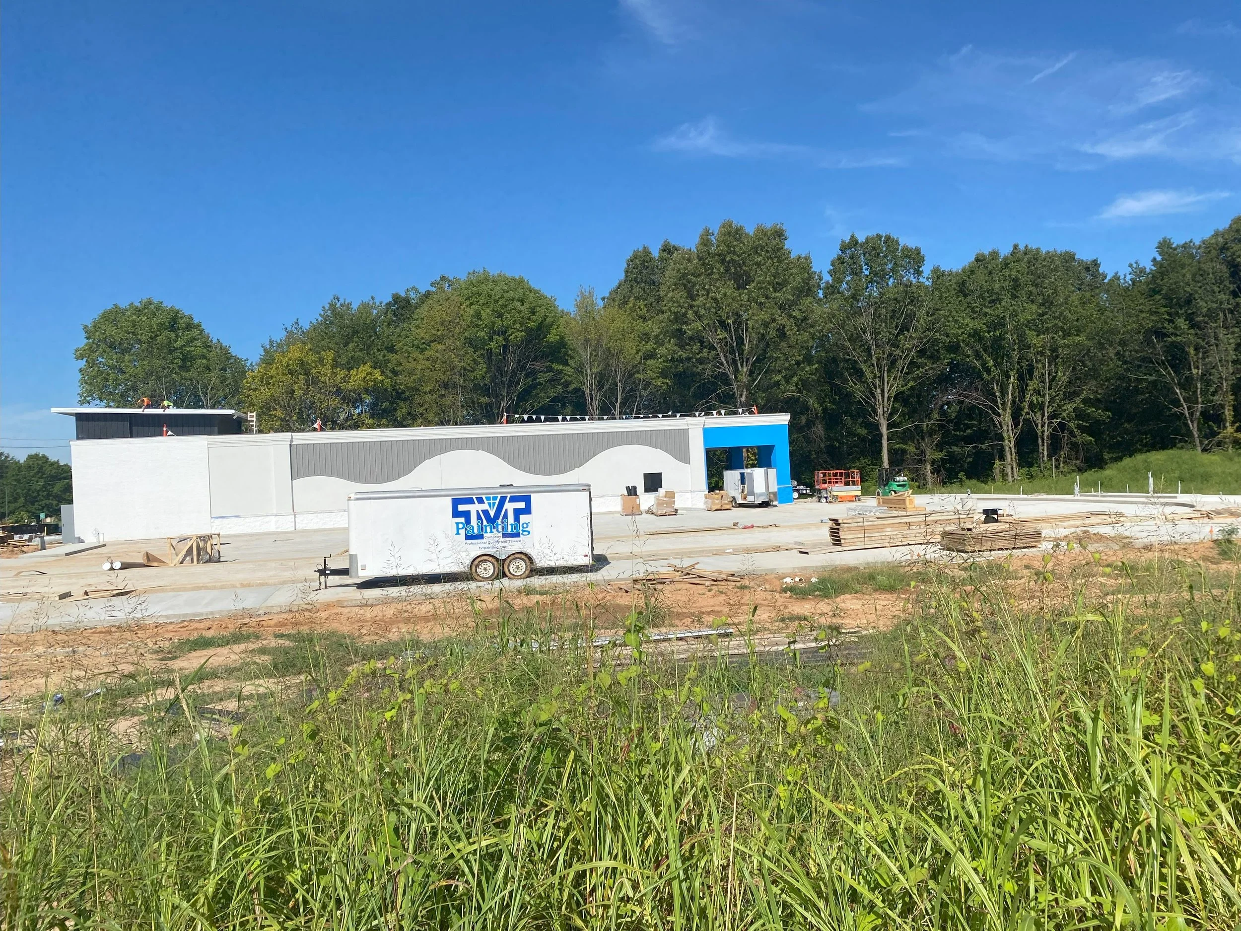Construction site with a white building, a trailer labeled 'Painting', construction materials, and a clear blue sky with trees in the background.