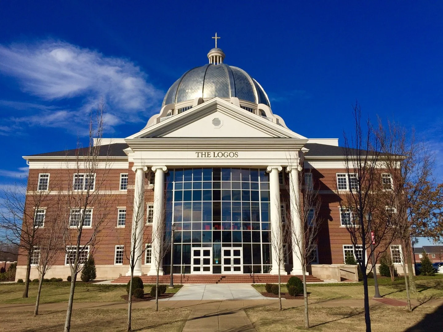 A large building with classical architecture, including white columns and a glass dome, labeled 'The Logos', under a blue sky with some clouds, and surrounded by leafless trees and grass.
