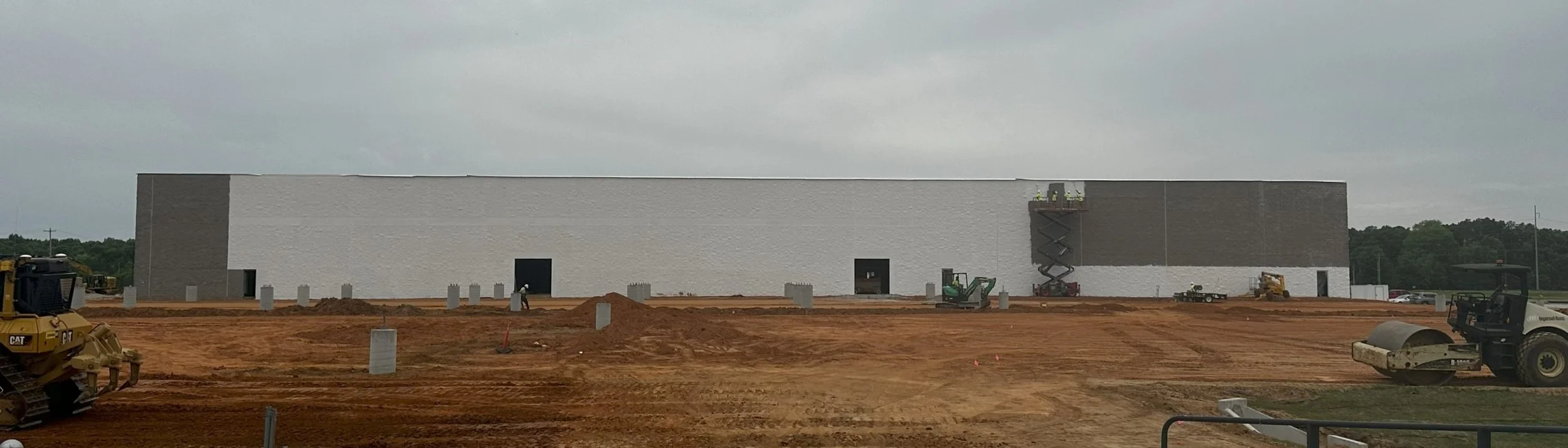 Construction site with a large, rectangular building under construction, with workers and construction vehicles, and a cloudy sky.