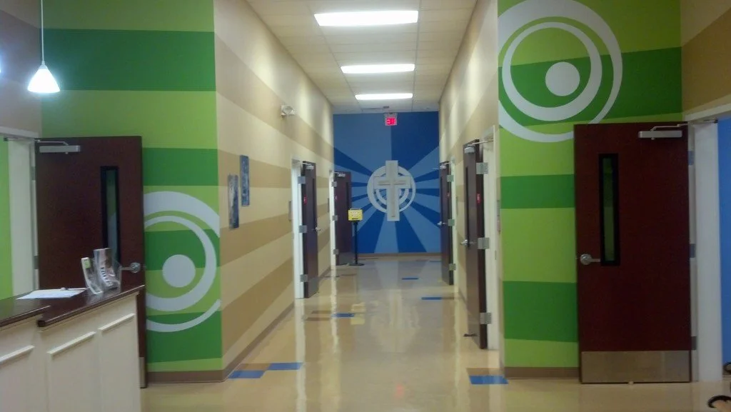 Hallway with green and beige striped walls, several dark brown doors with vertical window panels, and a blue mural with a white cross and radiating lines at the end of the corridor.