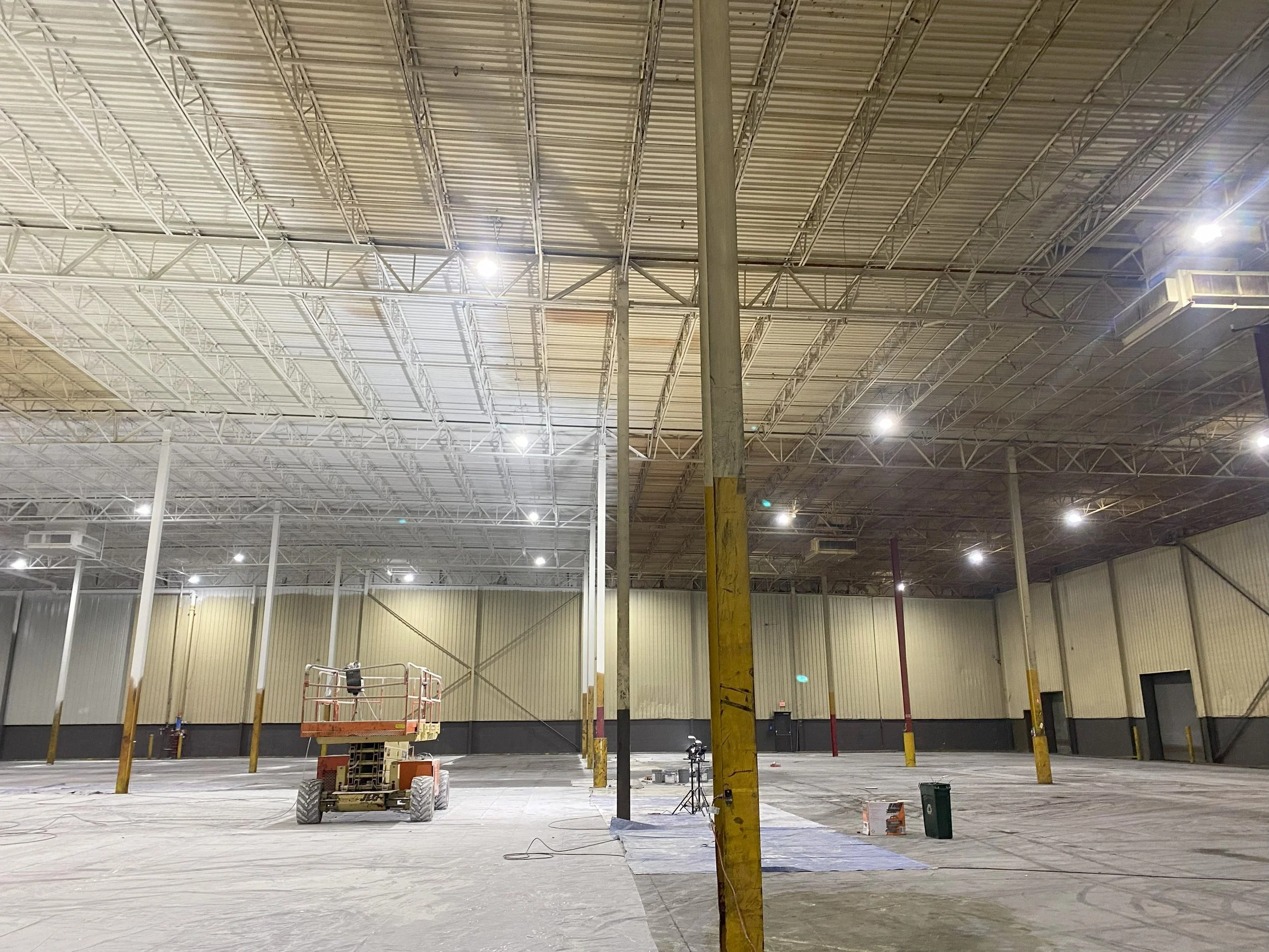 Interior of a large warehouse under construction, with exposed ceiling beams, steel columns, construction equipment, and scattered materials on the floor.