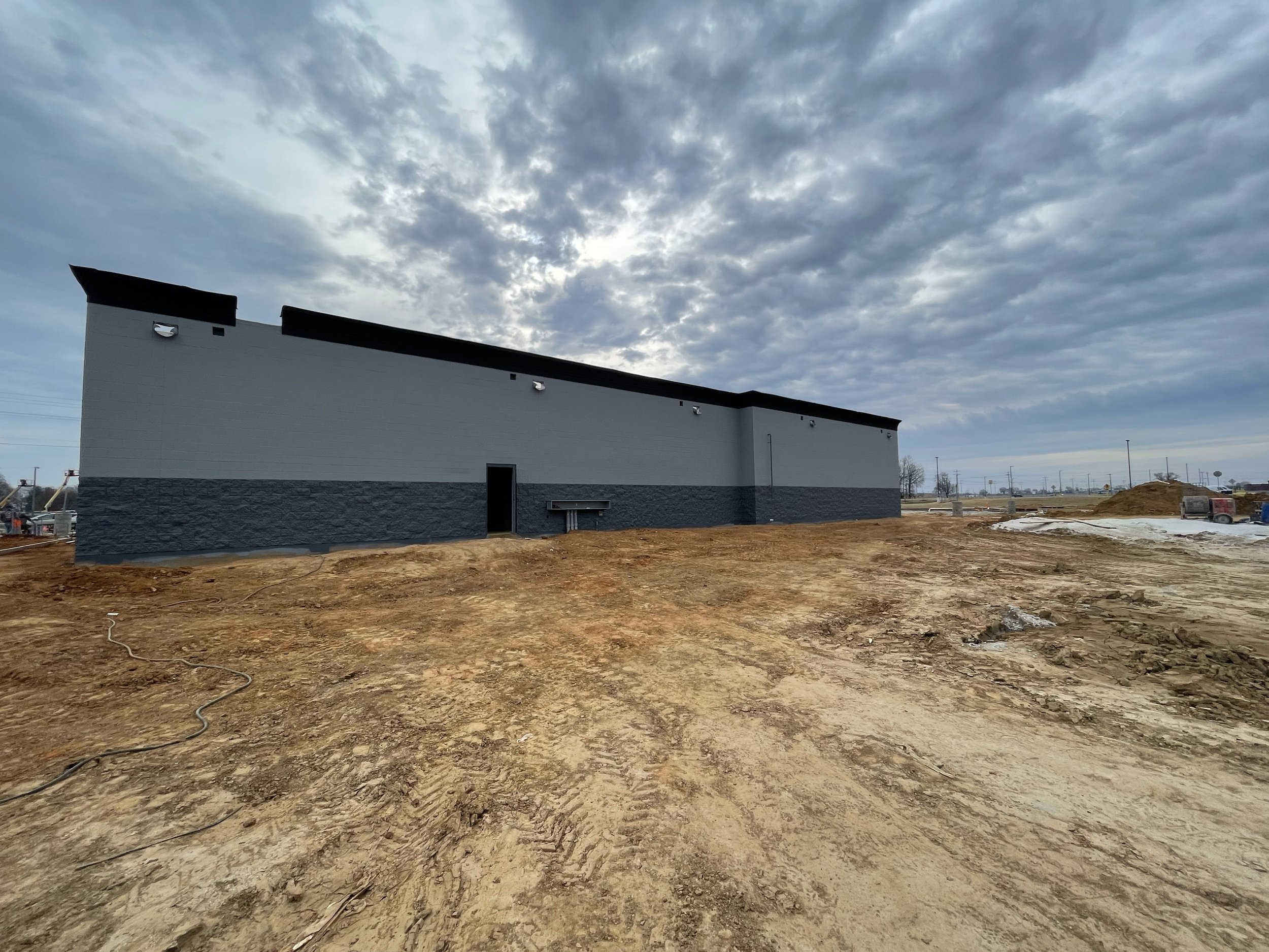 Construction site with dirt ground and a building with white and gray walls underneath a cloudy sky.