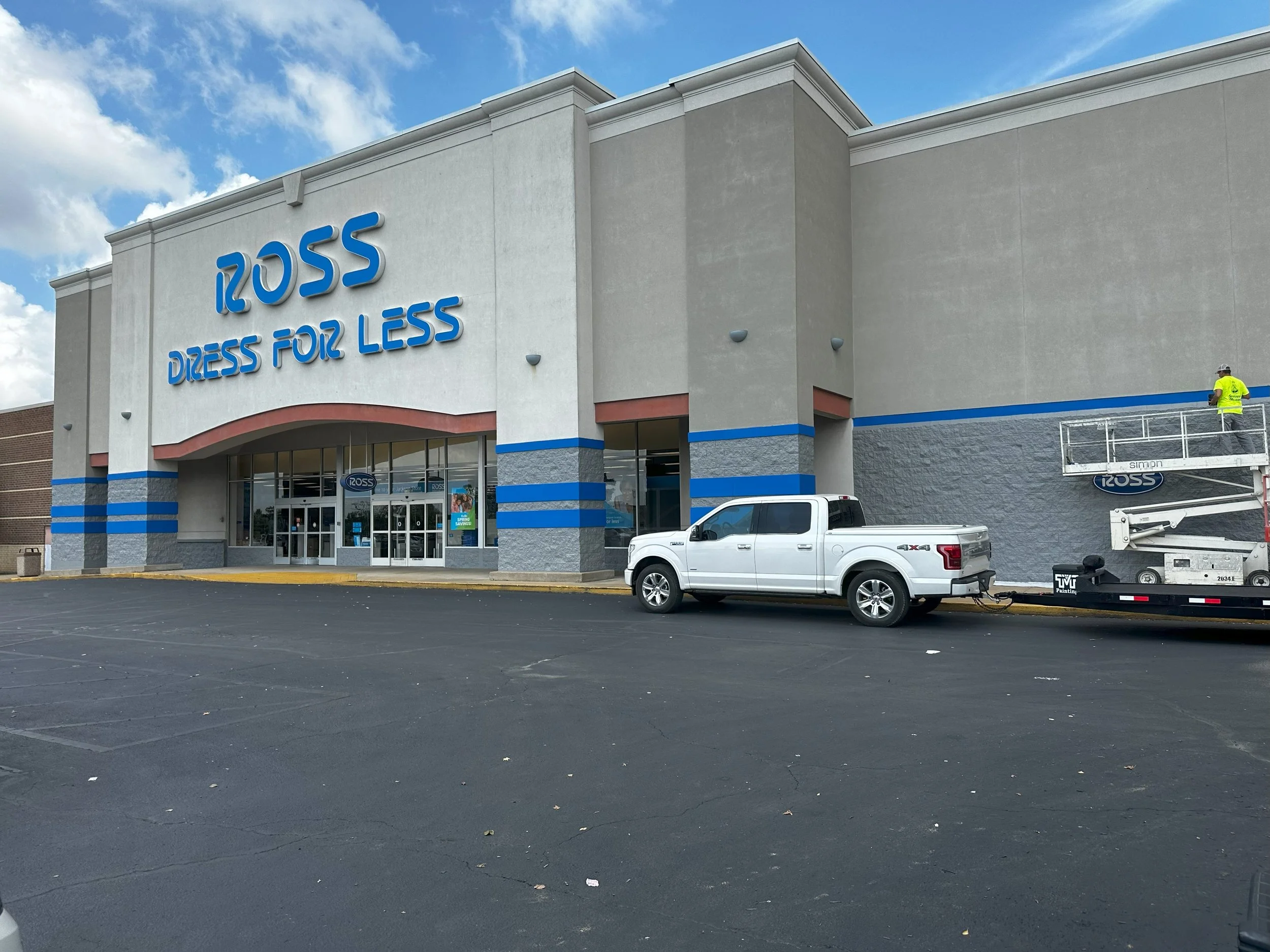 The exterior of a Ross Dress for Less store with a large sign on the facade, a parking lot with a white pickup truck and a utility lift with a worker, and partly cloudy sky.