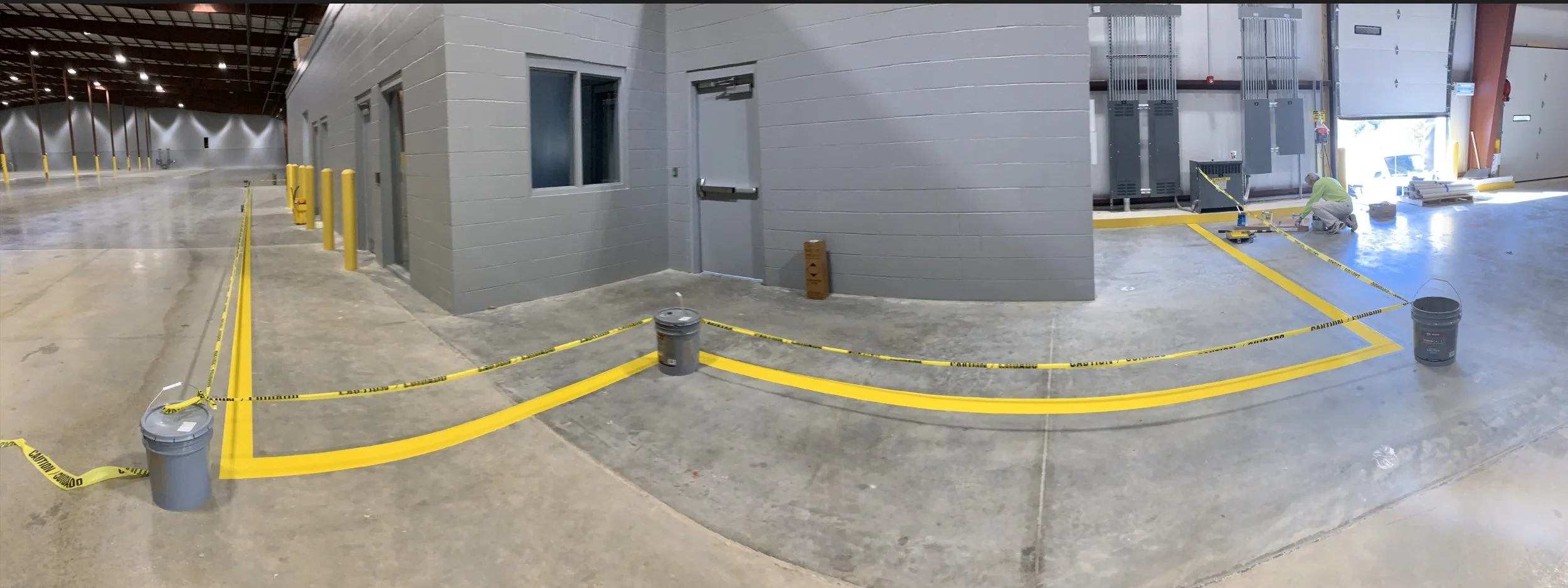 Interior view of a warehouse or industrial facility under construction or maintenance, with caution tape and buckets on the floor, and a worker installing or repairing equipment near a large open bay door.