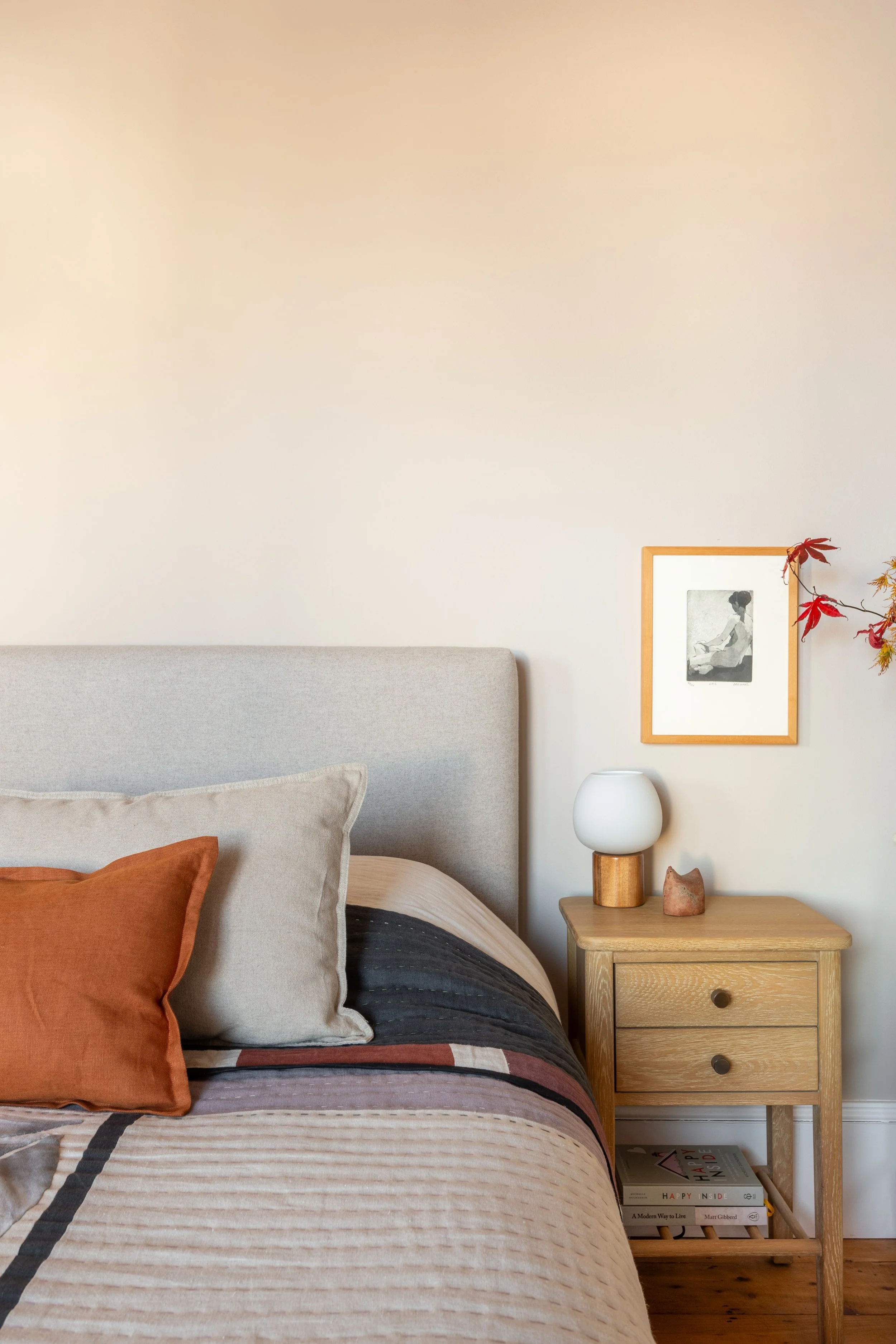 Close up of bed in warm neutral colours with cushions and handmade quilted bedspread, wooden bedside table and bedside light with wooden base and glass orb shade.