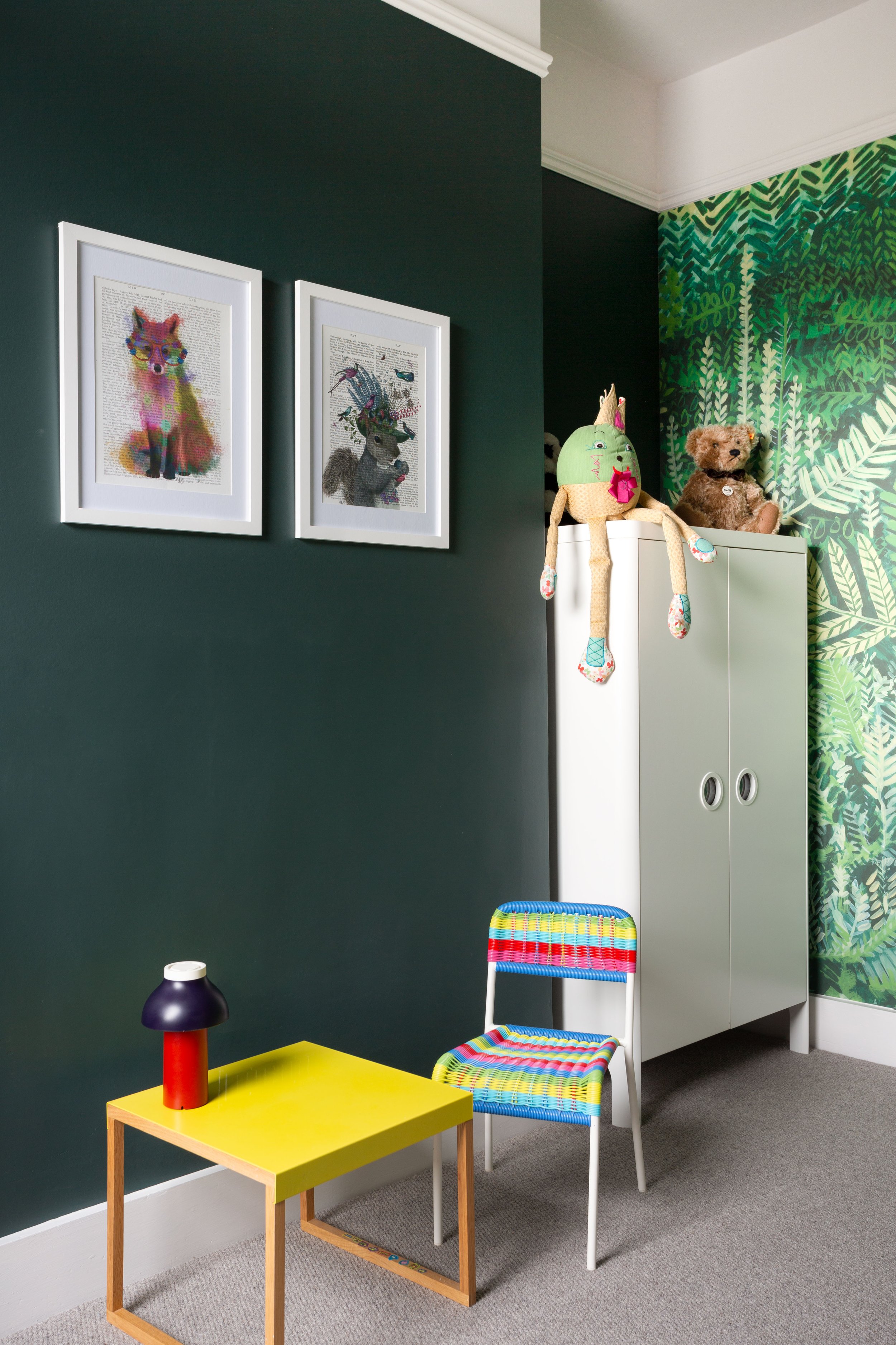 Child's bedroom with rich dark green wall, forest wall paper, contrasting white wardrobe, yellow side table, chair woven in rainbow colours and framed artwork.