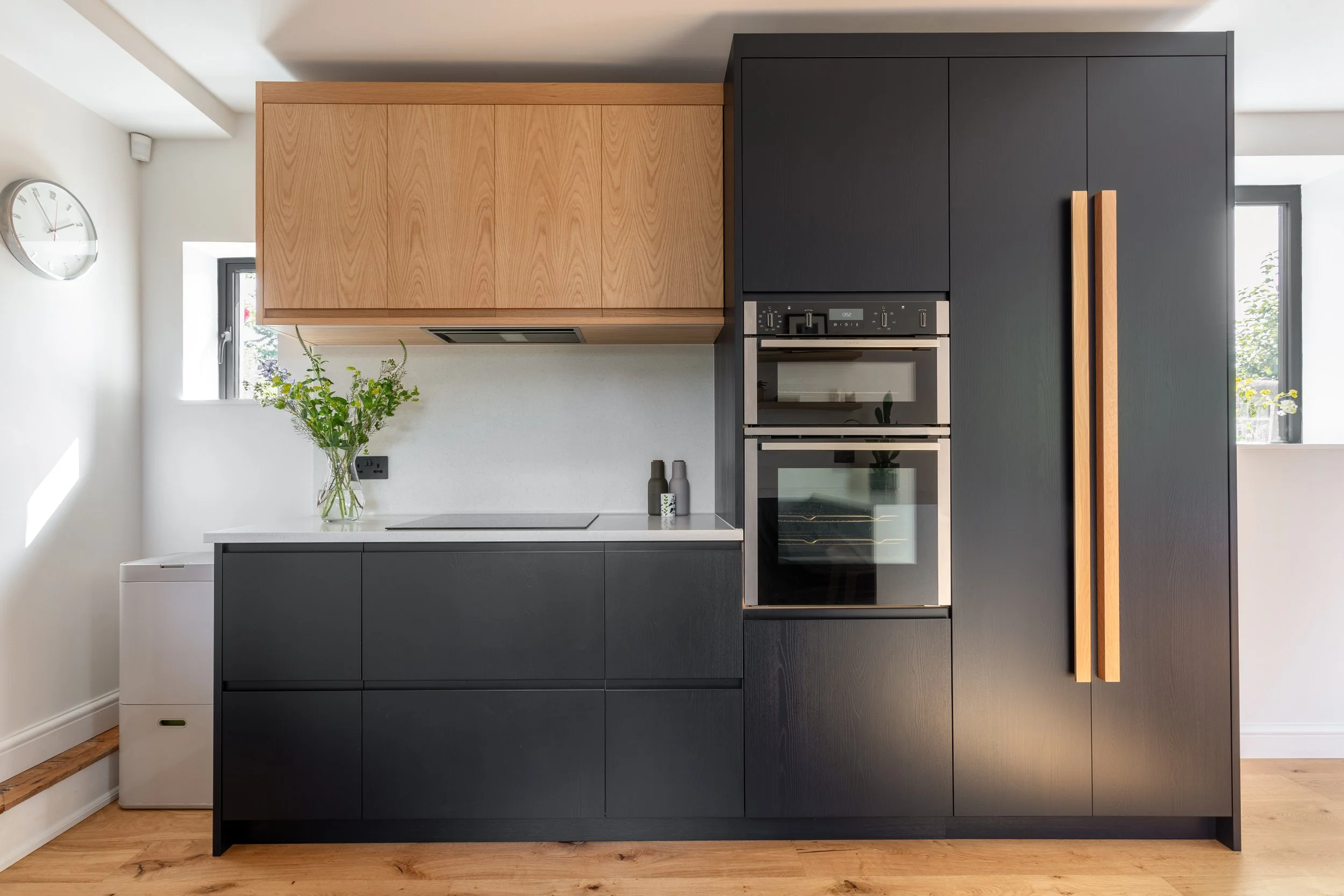 Contemporary kitchen with dark inky blue/black larder and base units contrasting with warm oak wall units, built-in oven, off white Wharf seamless countertop, a small window, and a glass vase with green plants on the counter.