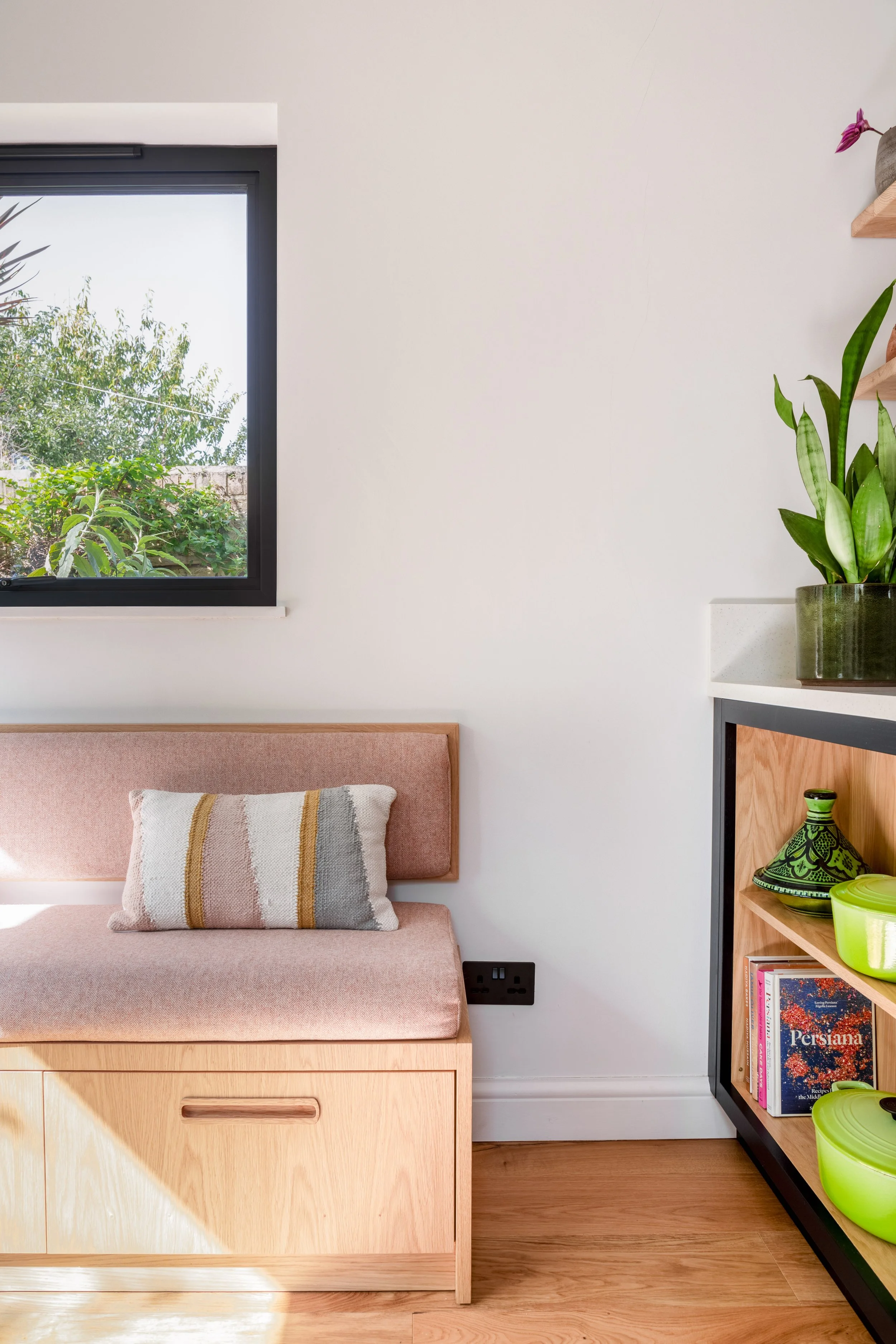 A cozy dining corner of a contemporary kitchen featuring an oak bench upholstered in pale terracotta fabric shelves built in to the back of the kitchen peninsula with green decor, a potted plant, and a large picture window showing greenery outside.