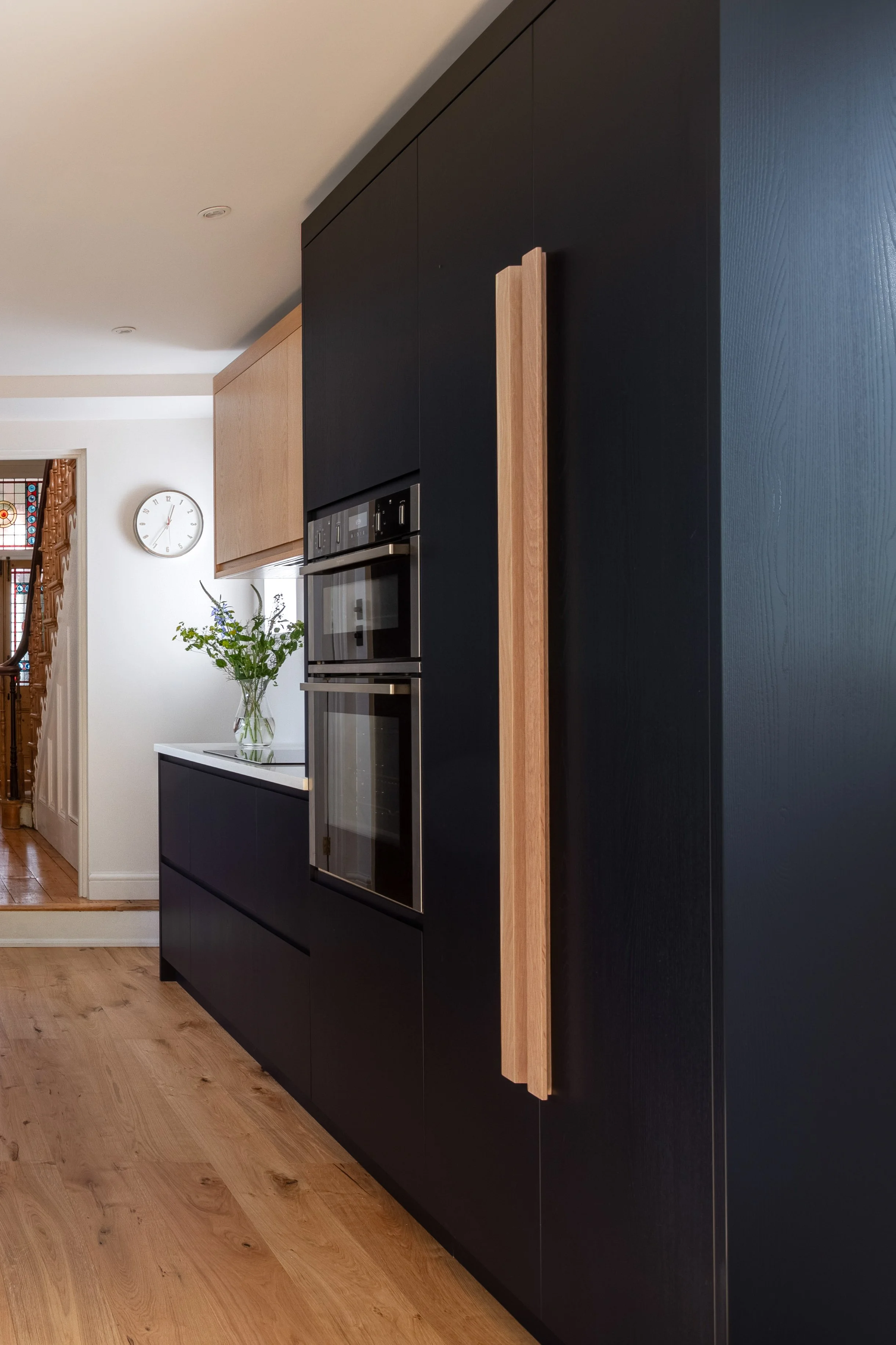 Contemporary kitchen featuring dark inky blue/black and oak wood cabinetry, full length wooden handles, built-in oven, and a vase with flowers on a white countertop.