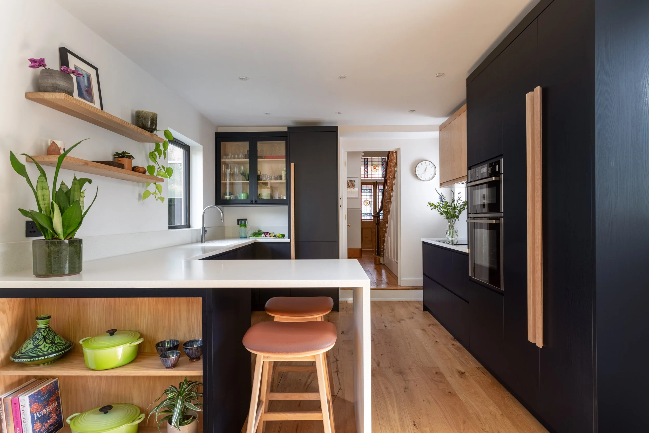 Modern kitchen with dark inky blue/black and contrasting oak cabinets, white countertops and styled open shelves and a kitchen peninsula with bar stools. The space leads to a hallway with a staircase and decorative stained glass window.