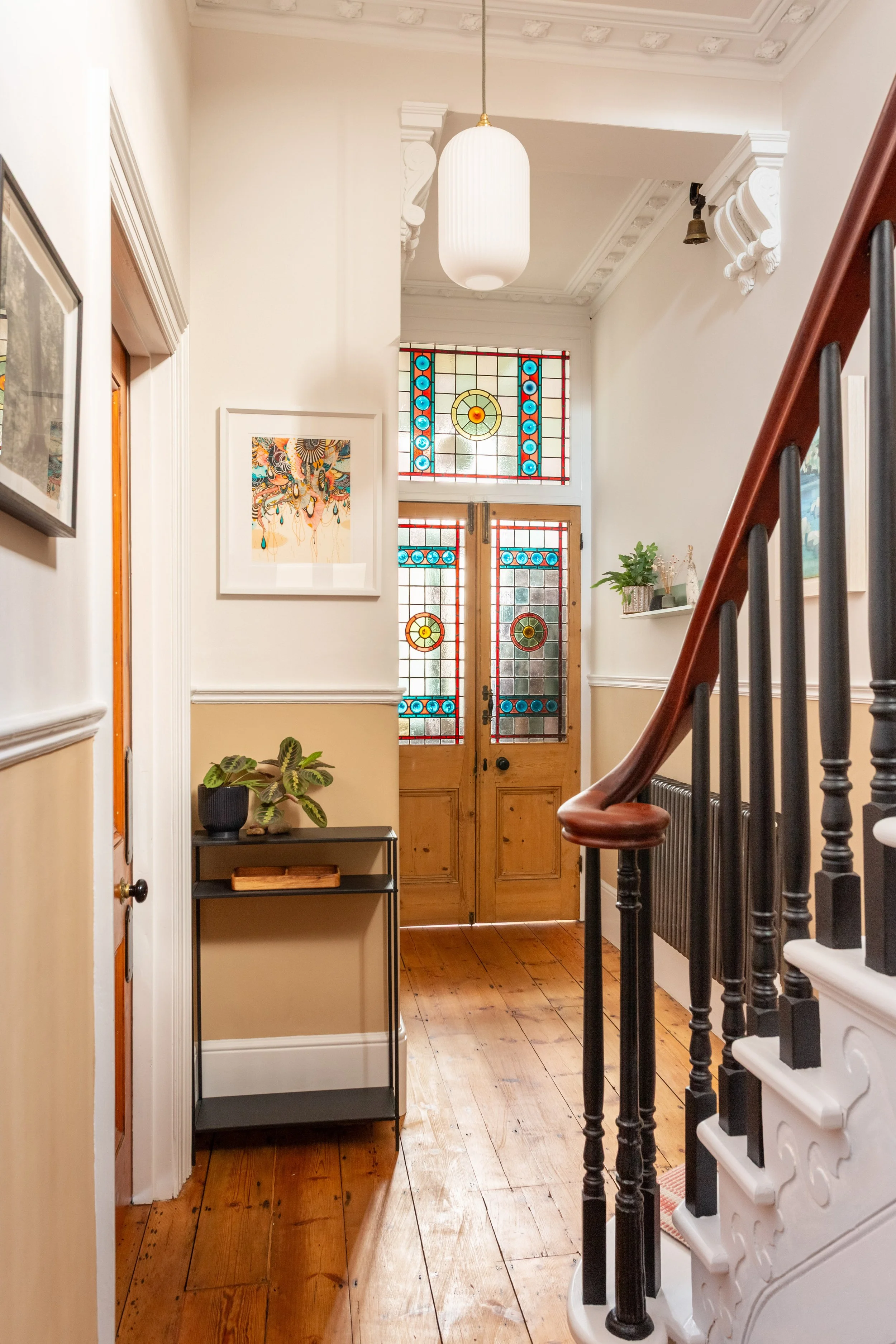 Beautiful wooden doors with stained glass lead to an entrance hall featuring a staircase with mahogany handrail, black spindles and white steps in a Victorian home. There are original wooden floors, a warm, neutral colour palette with bold accents.