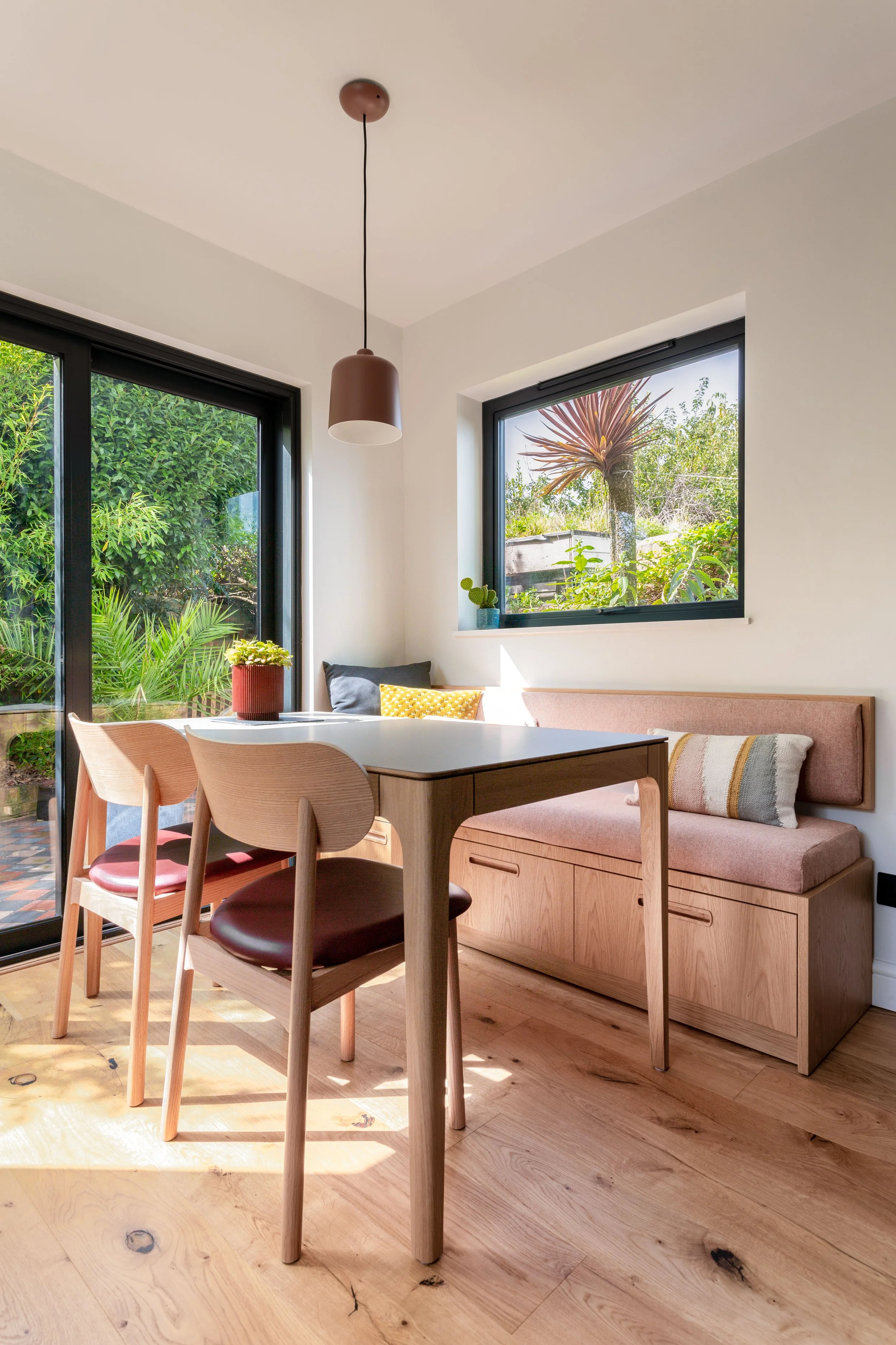 A cozy dining nook with oak bench finished with upholstered cushion, dining table with chairs , a large window and sliding door with greenery outside, potted plants on the windowsill, and a modern pendant lamp hanging from the ceiling.