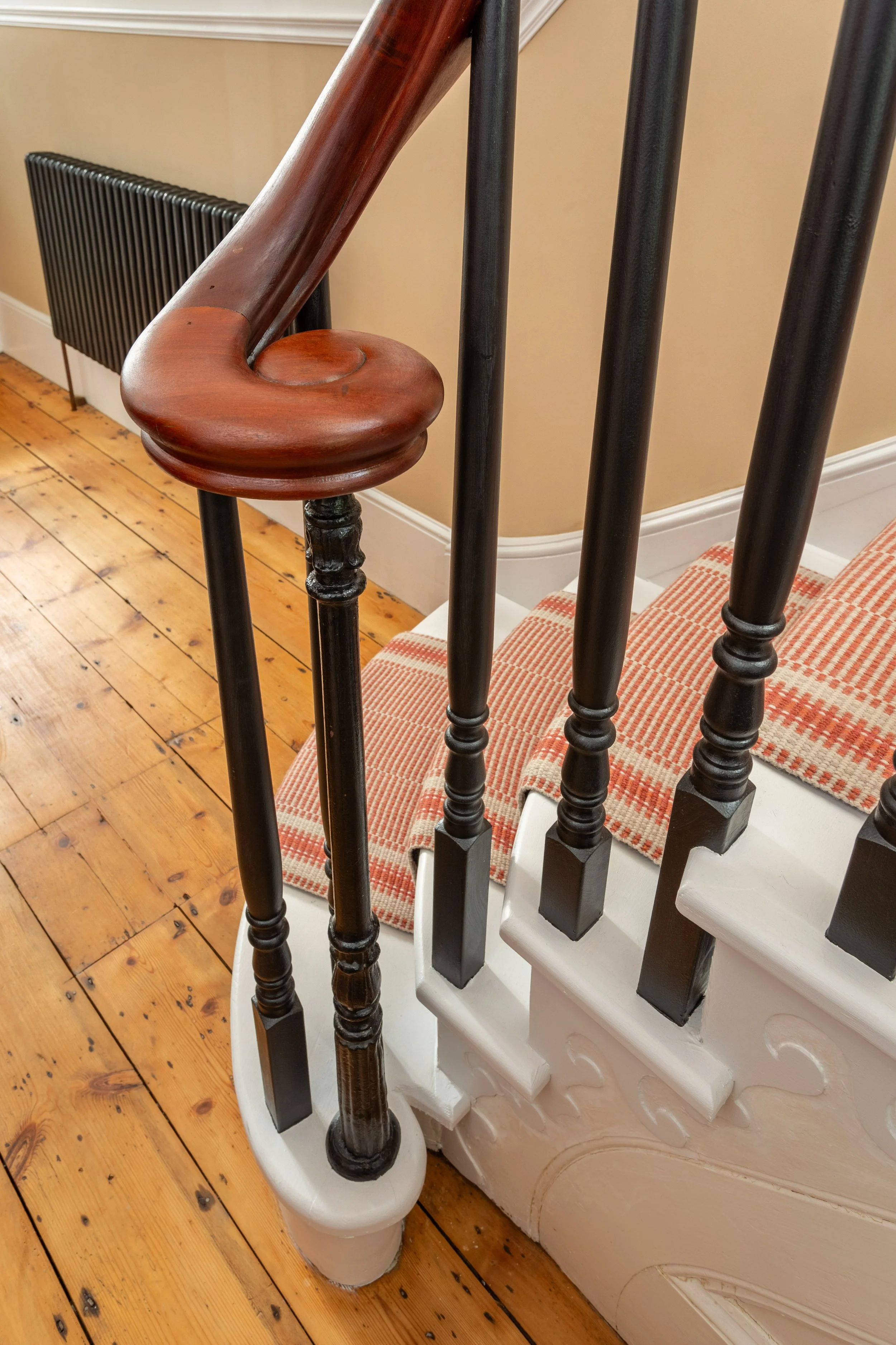 Close-up of an entrance hall staircase with mahogany handrail,  black spindles and white steps with a Roger Oates Mila terracotta runner. The hallway has original wooden flooring.