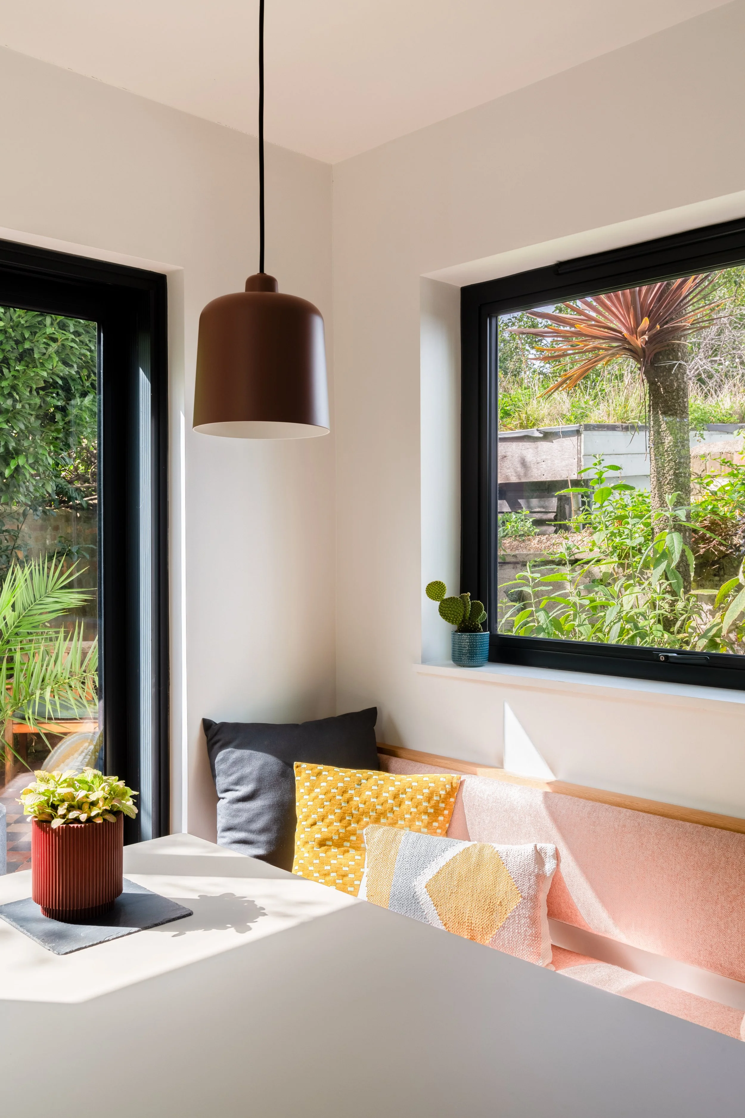 Bright dining corner of a contemporary kitchen with cosy bespoke banquette seating area, large windows showing greenery outside, a white table with a potted plant, and cushions on a seating area.