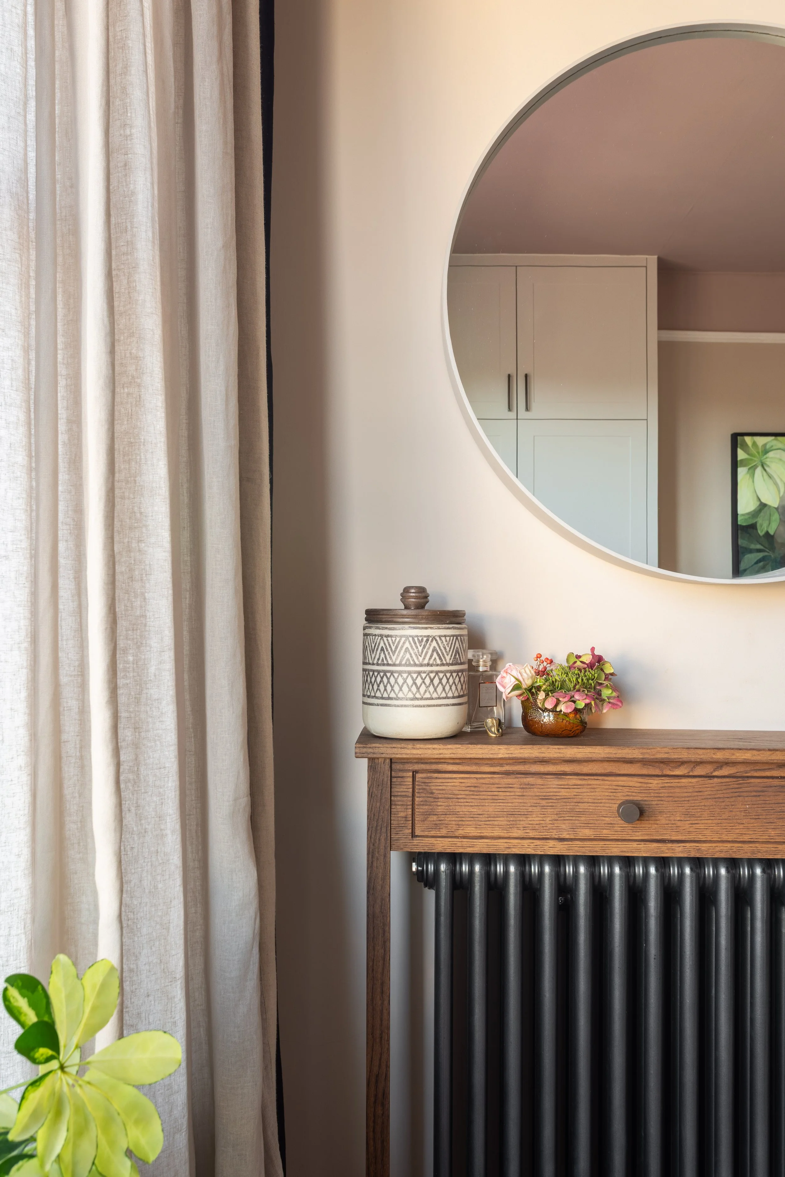 Close up of bedroom over radiator console table, with accessories, large round mirror overhead,  sheer full light curtains and green plant.