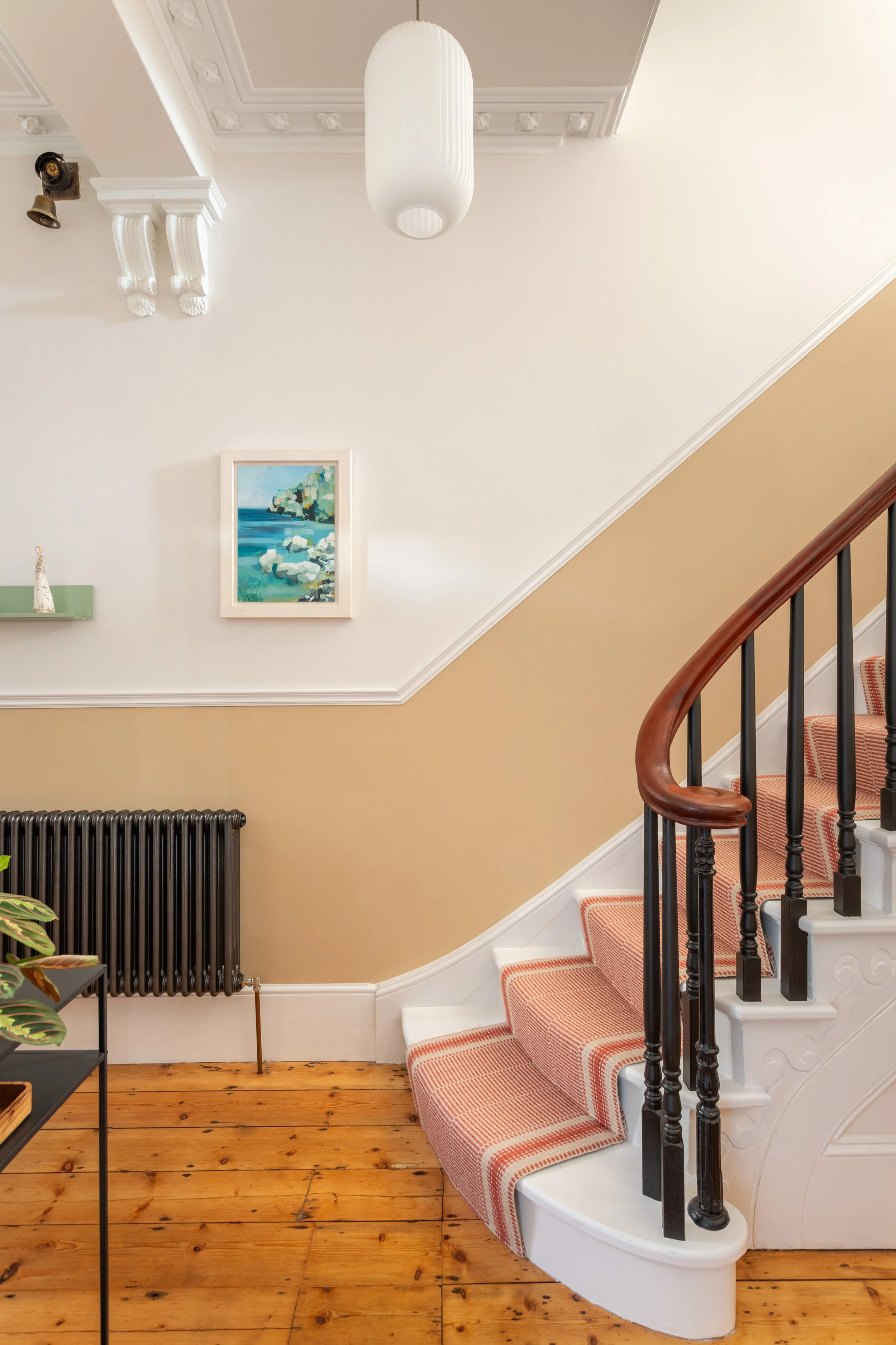 Entrance hall in a Victorian house showing a staircase with mahogany handrail,  black spindles  and white steps with a Roger Oates Mila terracotta runner. The ceiling features decorative moulding of the period and a white glass pendant light. 