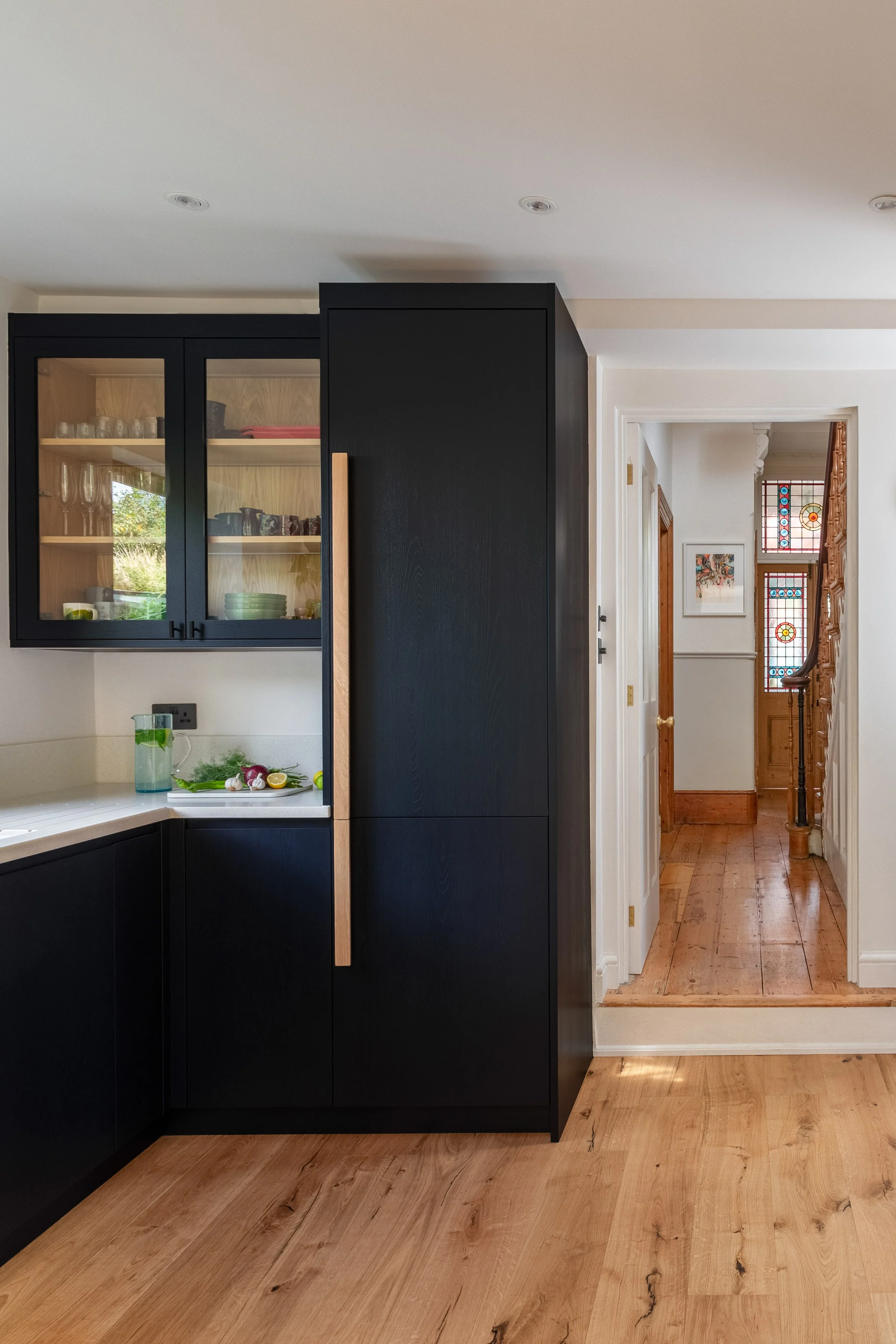Contemporary kitchen with ink blue/black and contrasting oak cabinetry, warm wood flooring. View to entrance hall with period stained glass windows.