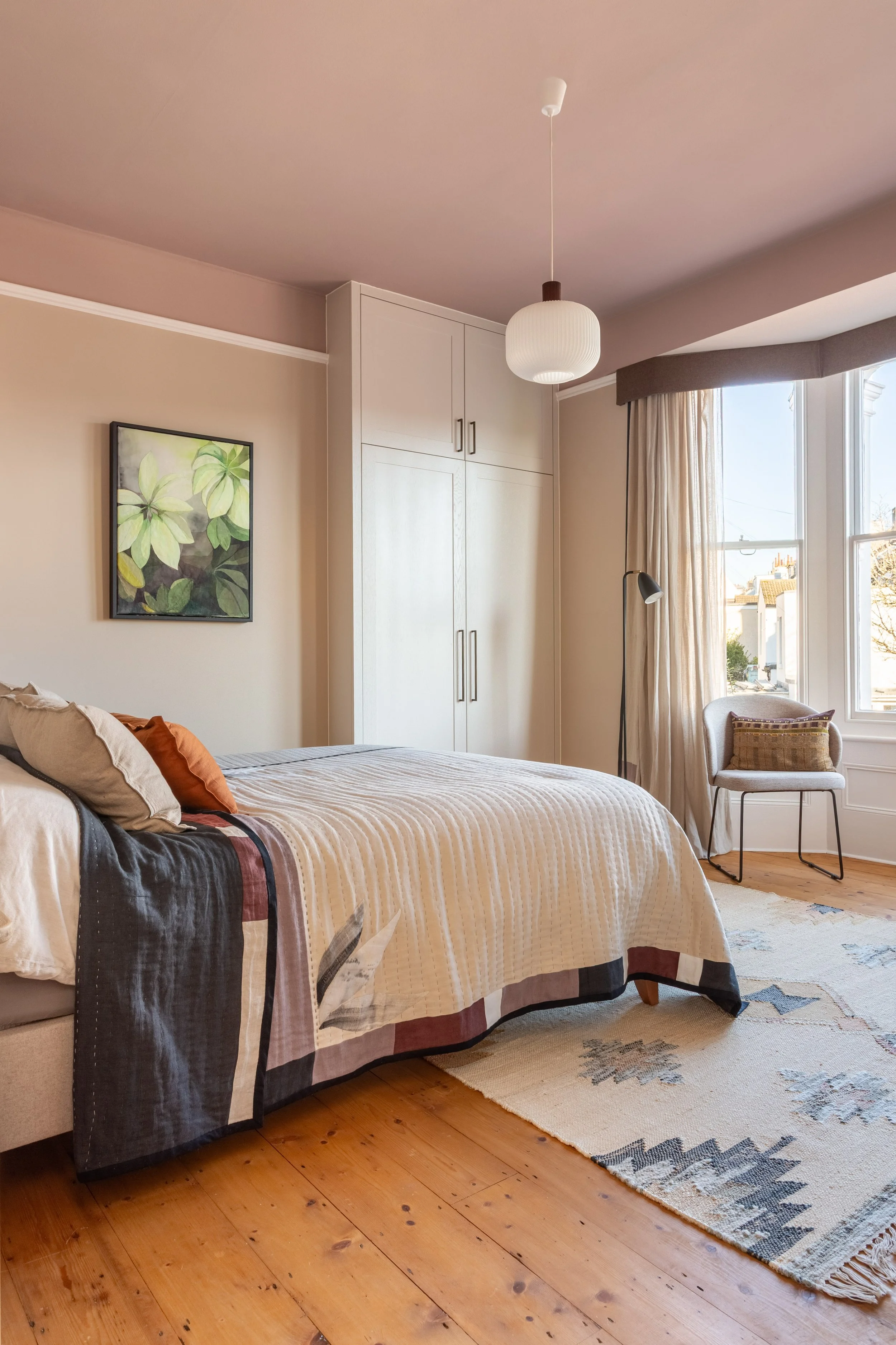 Elegant bedroom with warm neutral  colour palette, bespoke wardrobe, wooden flooring, natural rug, accent chair, bespoke artwork and handmade textiles.