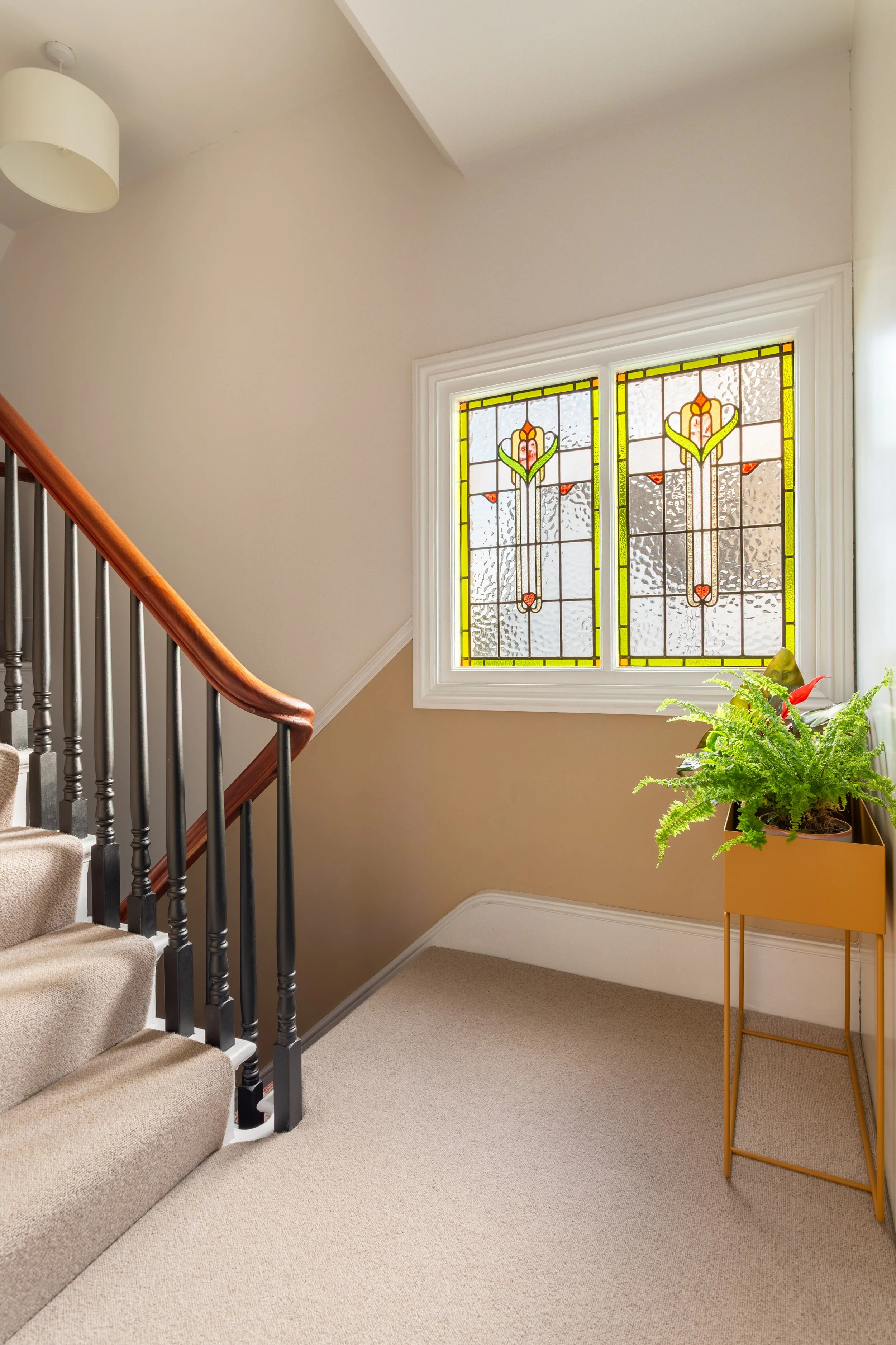 A staircase with carpeted steps and a wooden handrail, black spindles, a stained glass window in the wall on the landing  and an ochre Ferm Living planter with green plants.