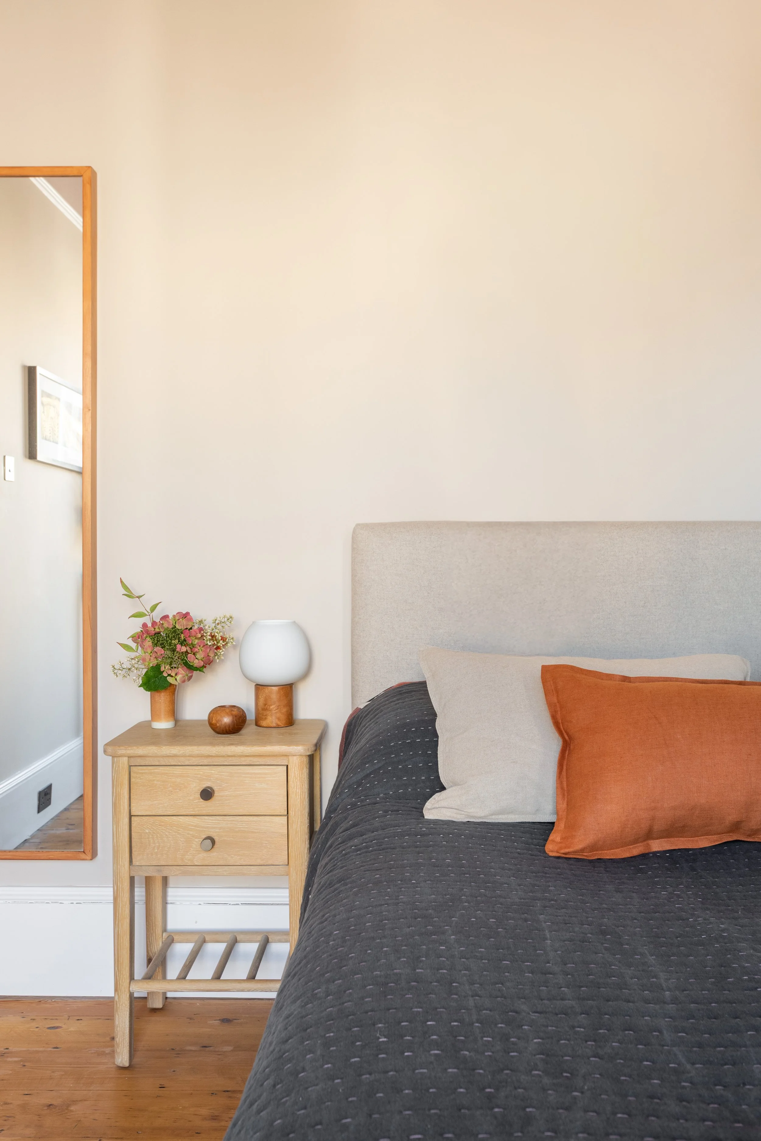 Close up of bed in warm neutral colours with cushions and handmade quilted bedspread, wooden bedside table and bedside light with wooden base and glass orb shade.