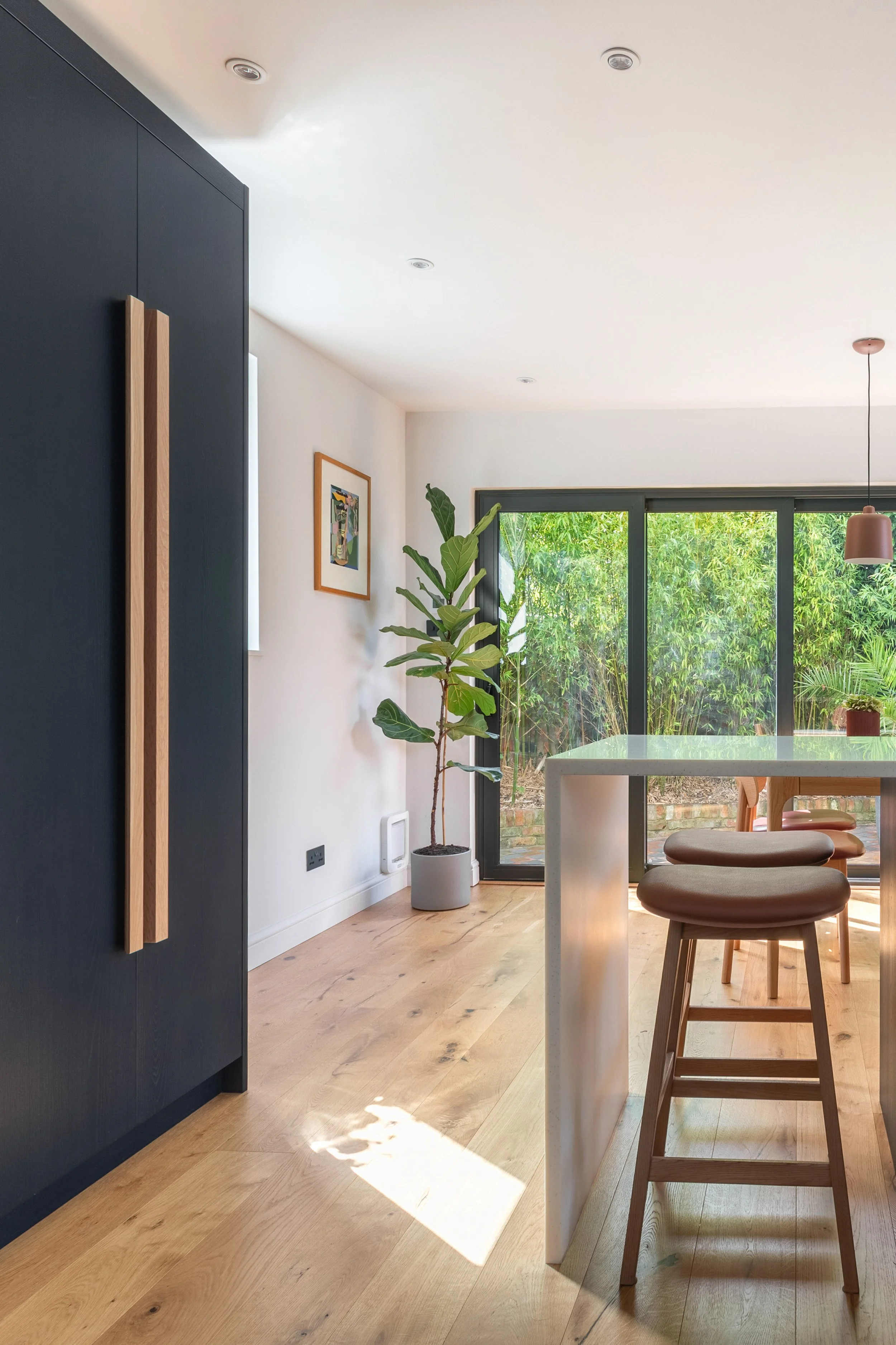 Contemporary kitchen with inky blue/black larder cupboard, an off white peninsula and wooden bar stools, large sliding glass doors with greenery outside, potted plant, framed artwork on wall, and engineered oak wooden flooring.