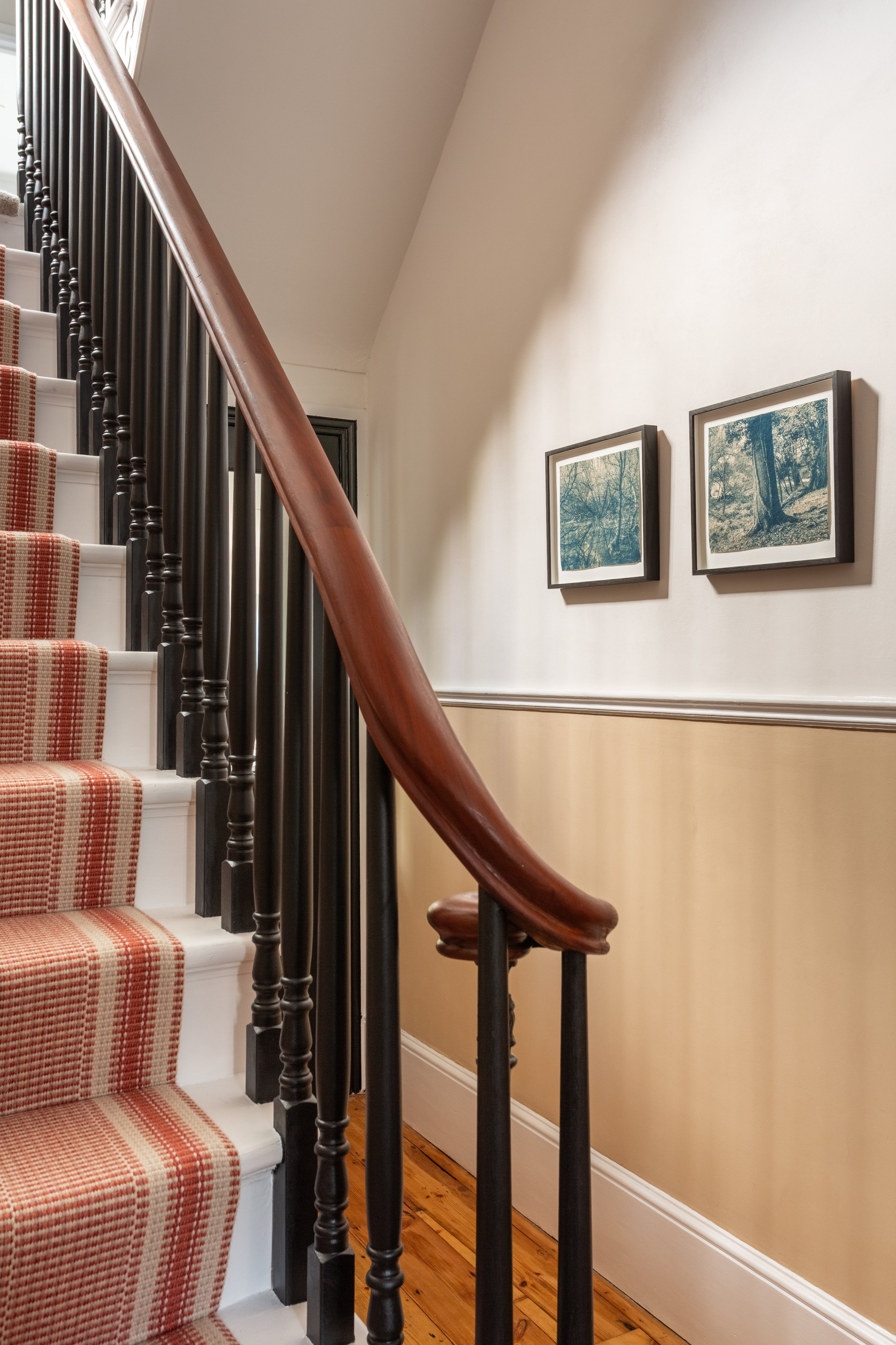 An entrance hall staircase with mahogany handrail,  black spindles and white steps with a Roger Oates Mila terracotta runner.  There is framed photography artwork of trees hanging on a wall with warm sand colour under the dado rail.