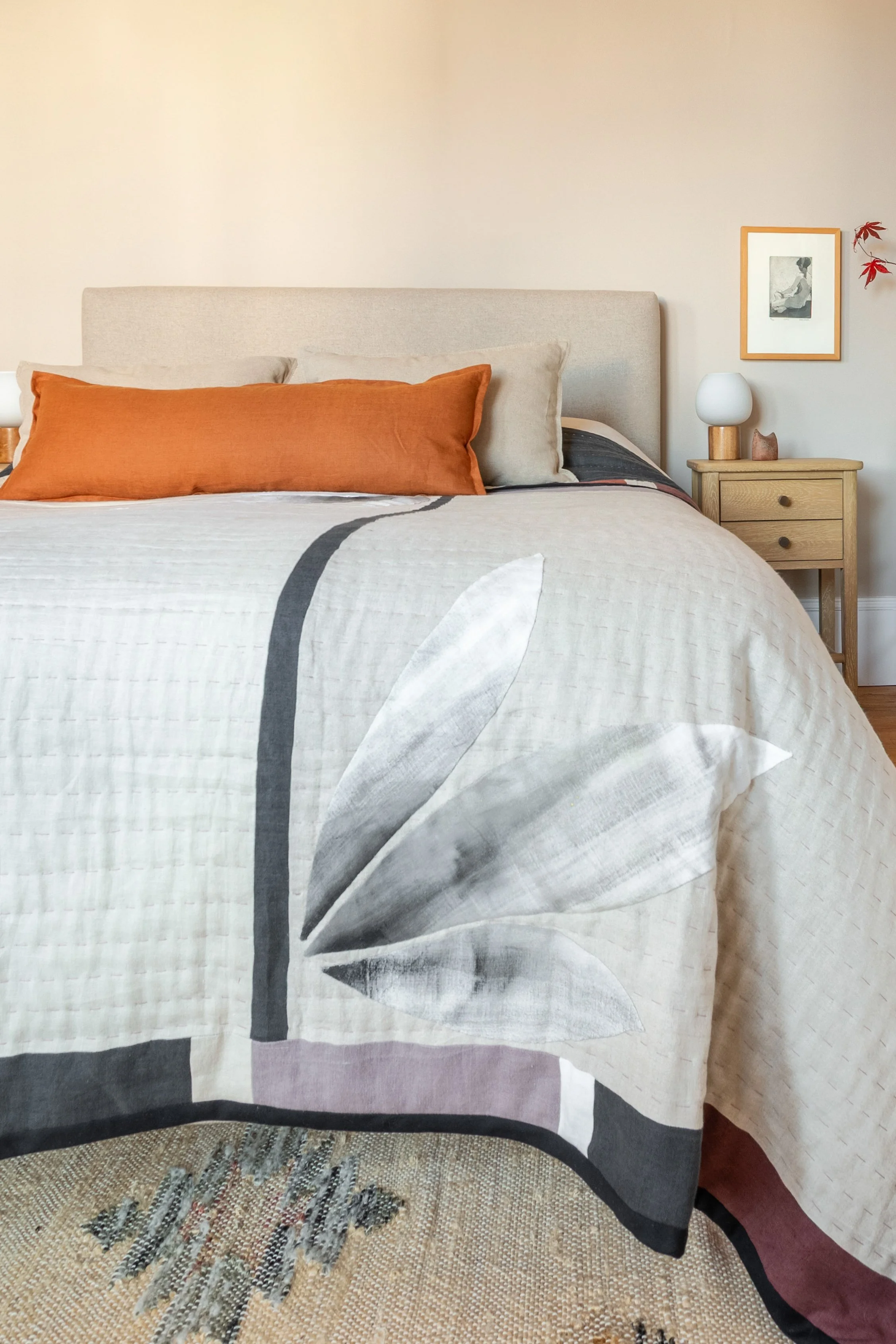 lose up of bed in warm neutral colours with cushions and handmade quilted and appliquéd linen bedspread, wooden bedside table and bedside light with wooden base and glass orb shade.