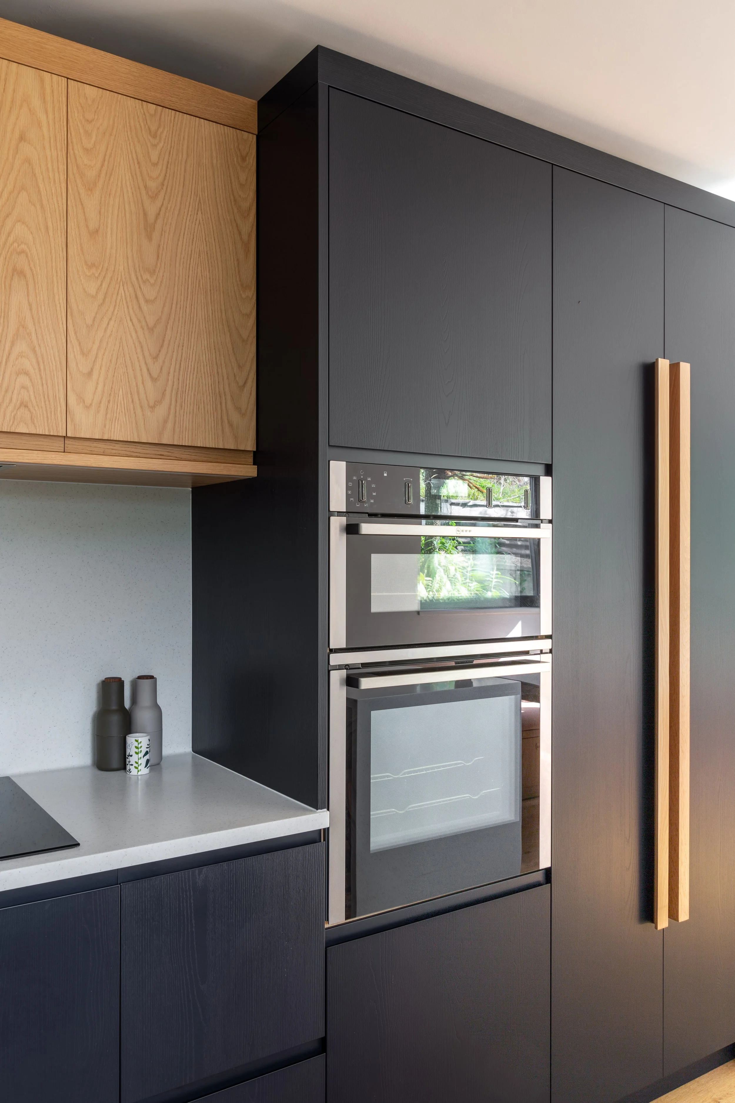 Close-up of modern kitchen with dark inky blue/black cabinetry, built-in stainless steel and black oven, oak wood cabinet accents, and decorative accessories on the countertop.