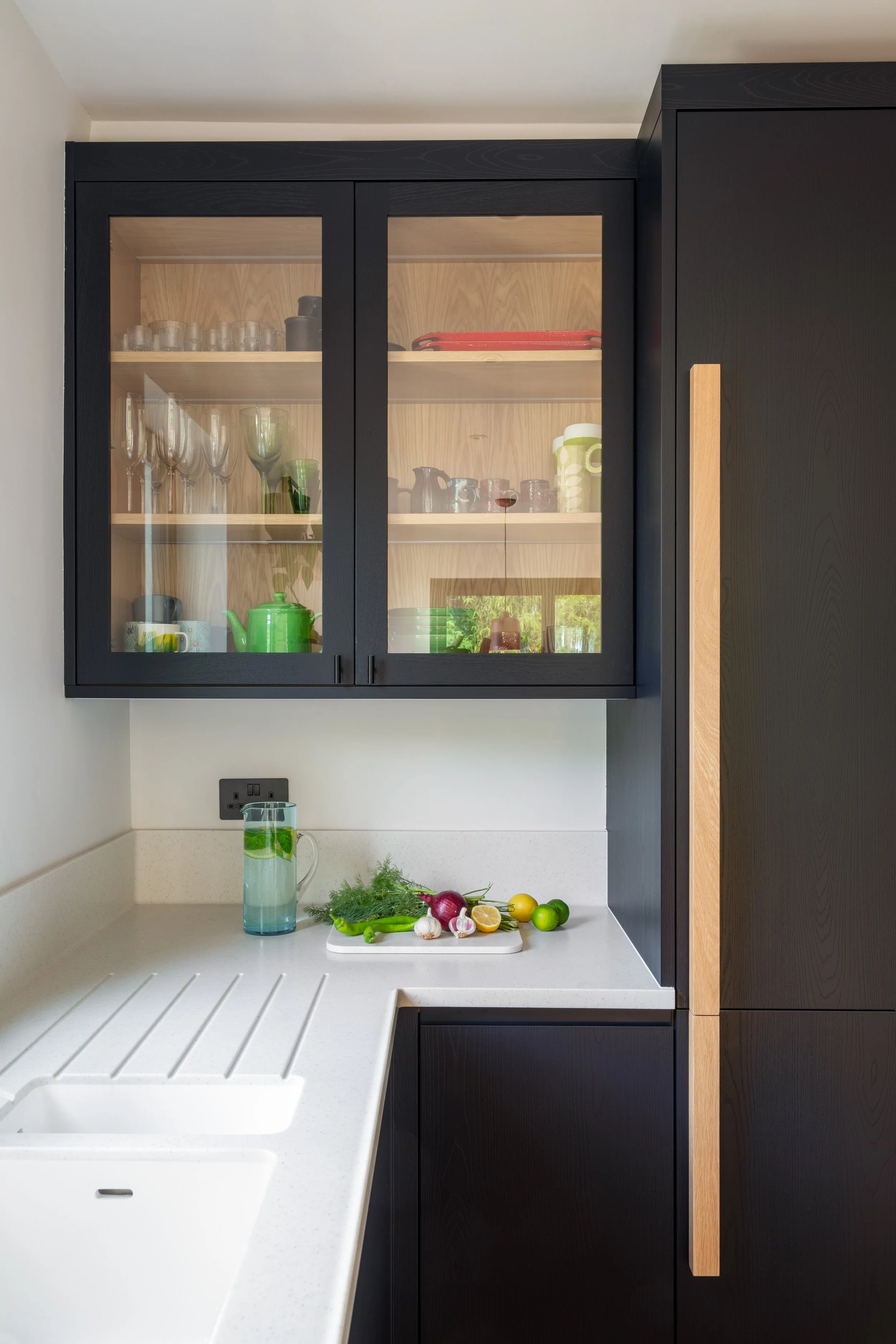 Wharf seamless kitchen worktop and a dark inky blue/black wall cabinet with glass panelled doors revealing oak shelves holding glasses, cups, and a green teapot.