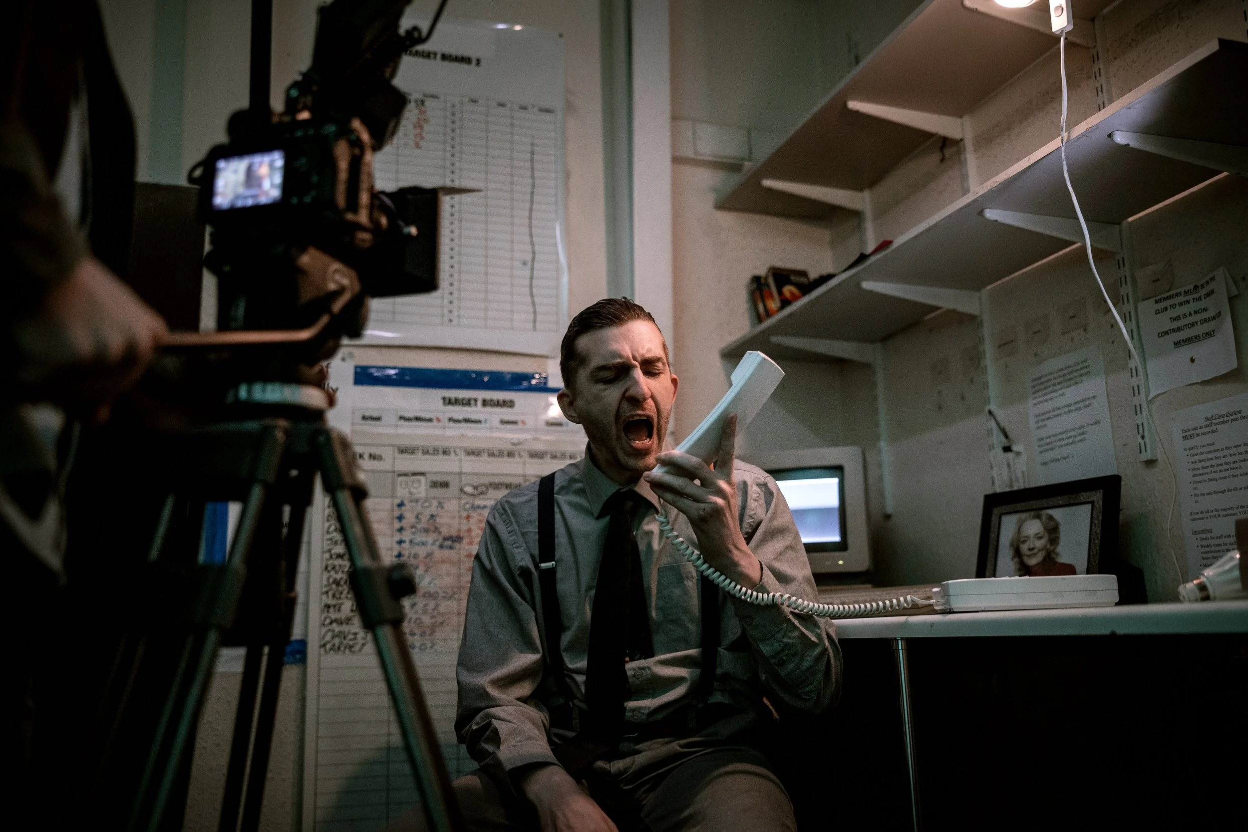 A man in a grey shirt and braces is sitting on the floor, yelling into a white telephone receiver, with a camera recording him. The scene of an office with shelves, a framed photo of a woman, and a target board on the wall.