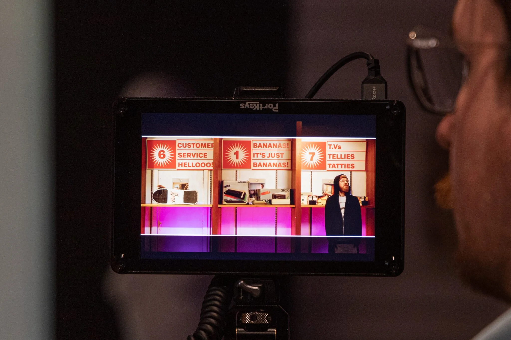 View of a video monitor showing three booths with bright red signs, each displaying text and a man with glasses and a black hoodie standing in front of the rightmost booth.