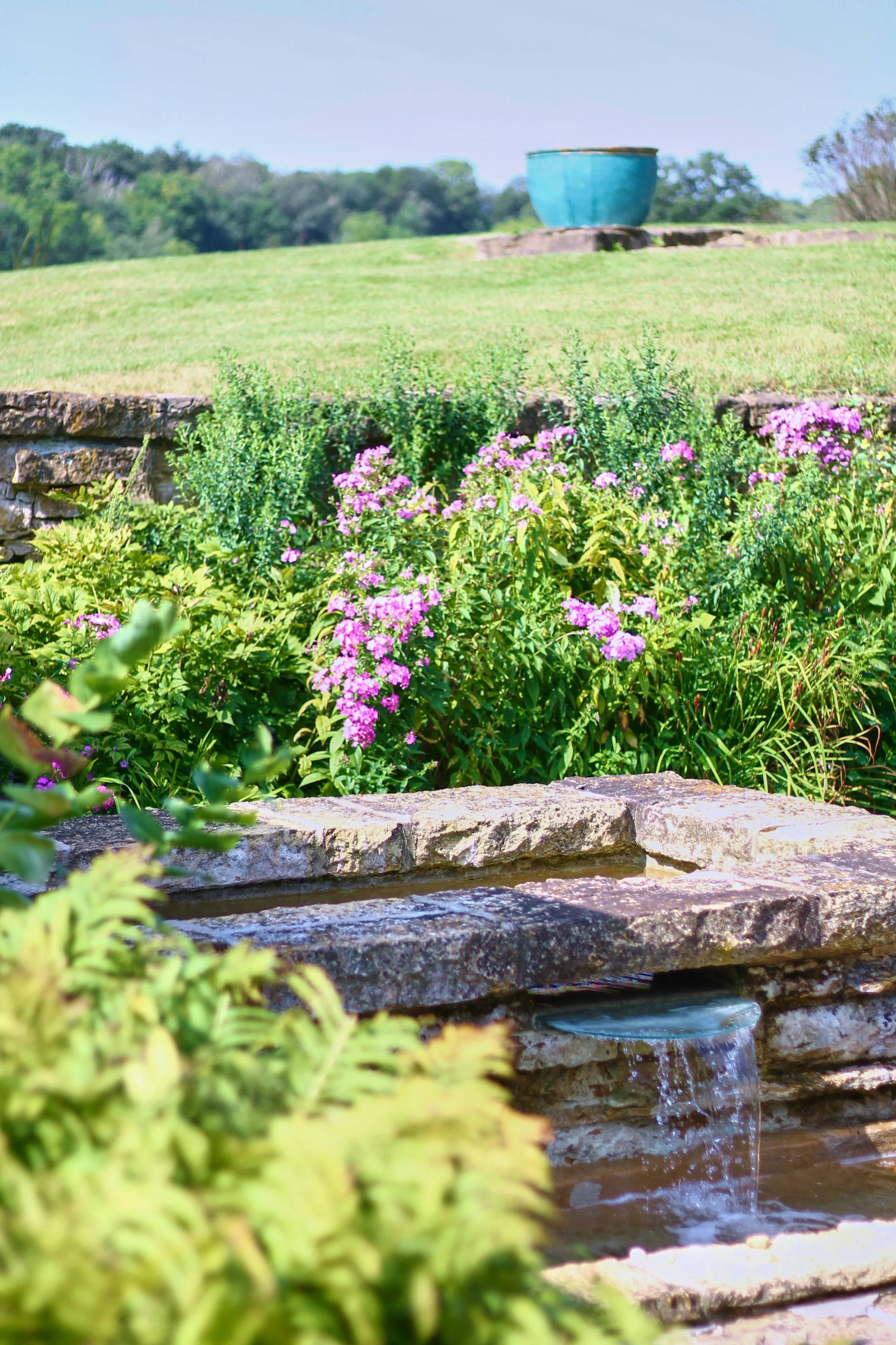 Sunlit landscaped garden featuring a natural stone fountain, flowing water, blooming purple flowers, and manicured greenery on a gently rolling lawn.